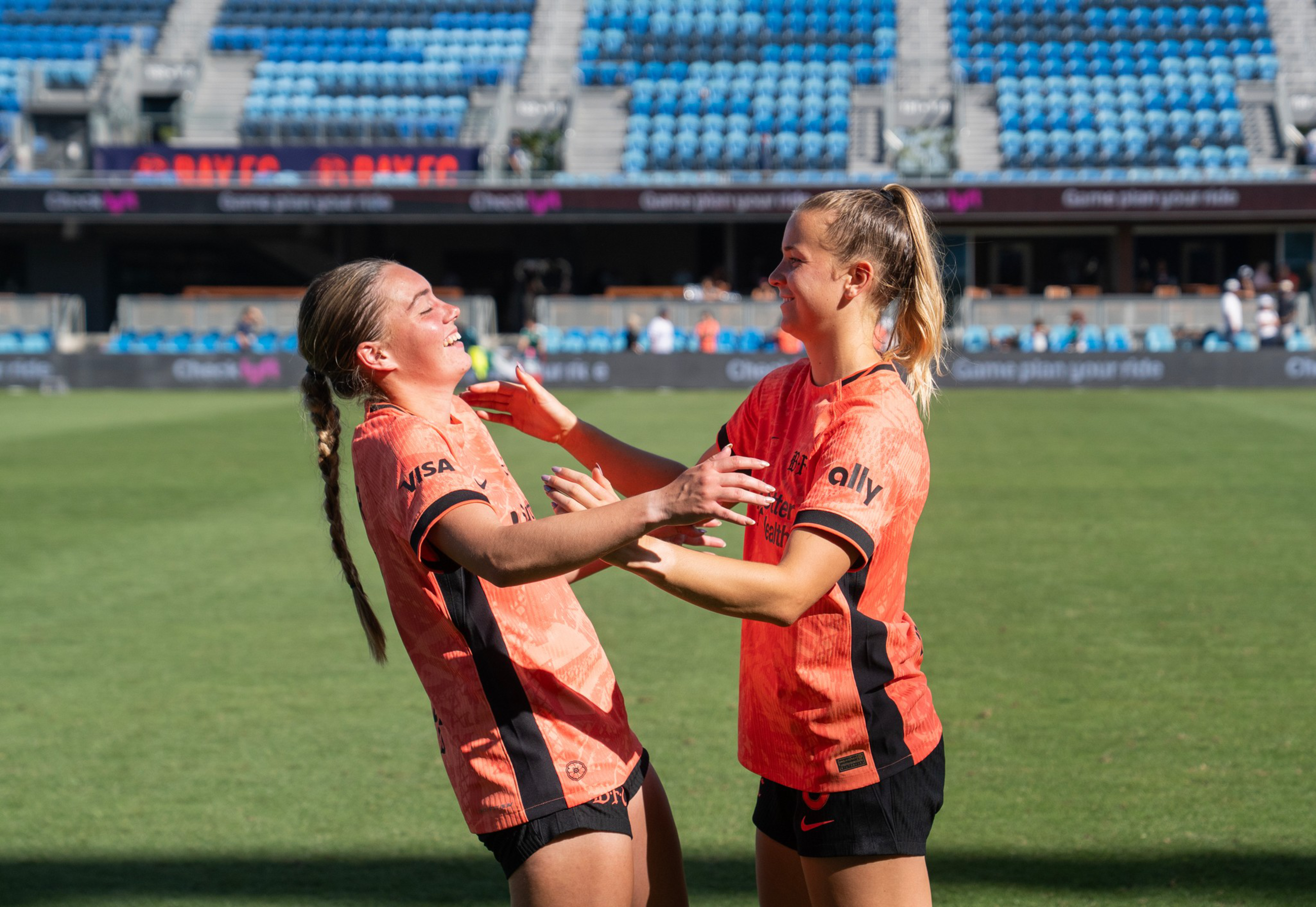 Two female soccer players in orange and black uniforms smile and reach out to embrace each other on a sunny field with empty blue stadium seats in the background.