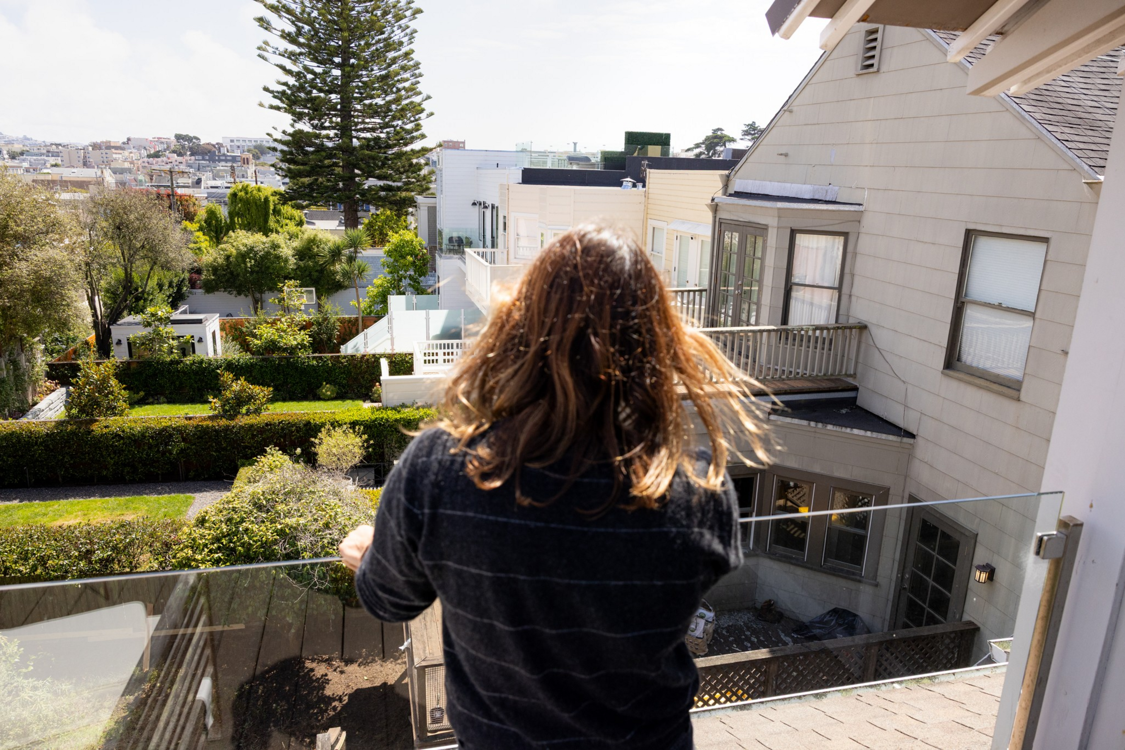 A person with long hair stands on a balcony overlooking a backyard, neighboring houses, and greenery under a bright sky.