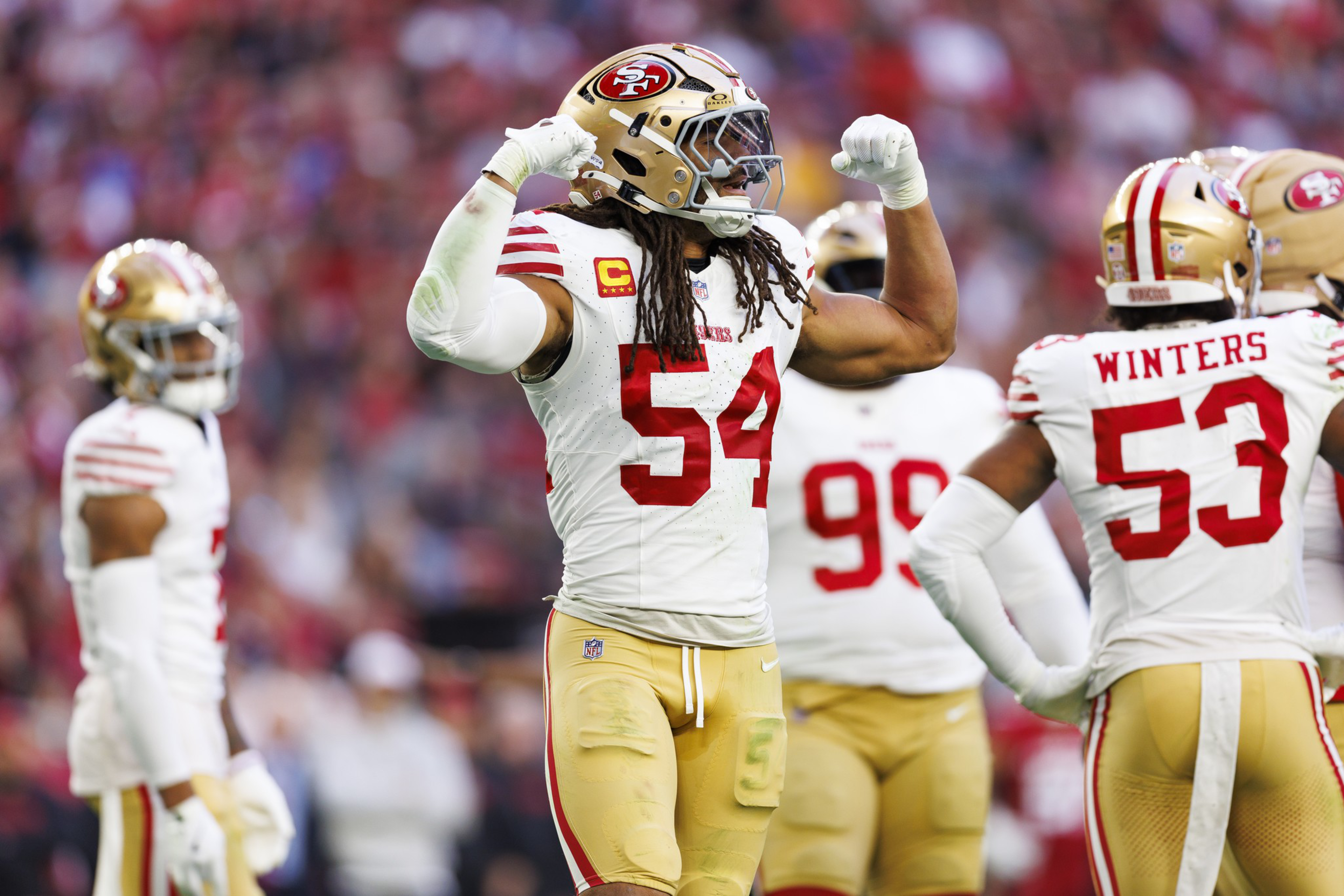A football player in a white and red uniform flexes his muscles on the field, surrounded by teammates wearing similar gear, with a blurred crowd in the background.