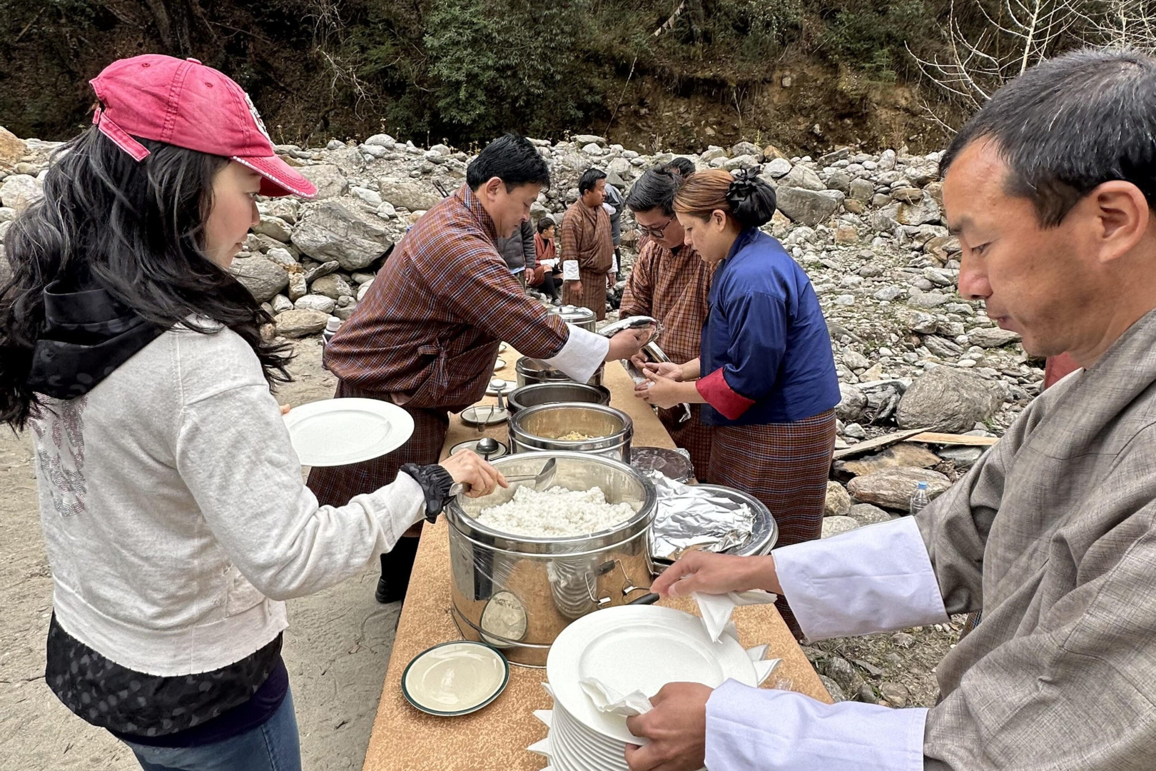 People dressed in traditional clothes and casual wear serve and receive food outdoors from large metal containers on a table by rocky terrain.