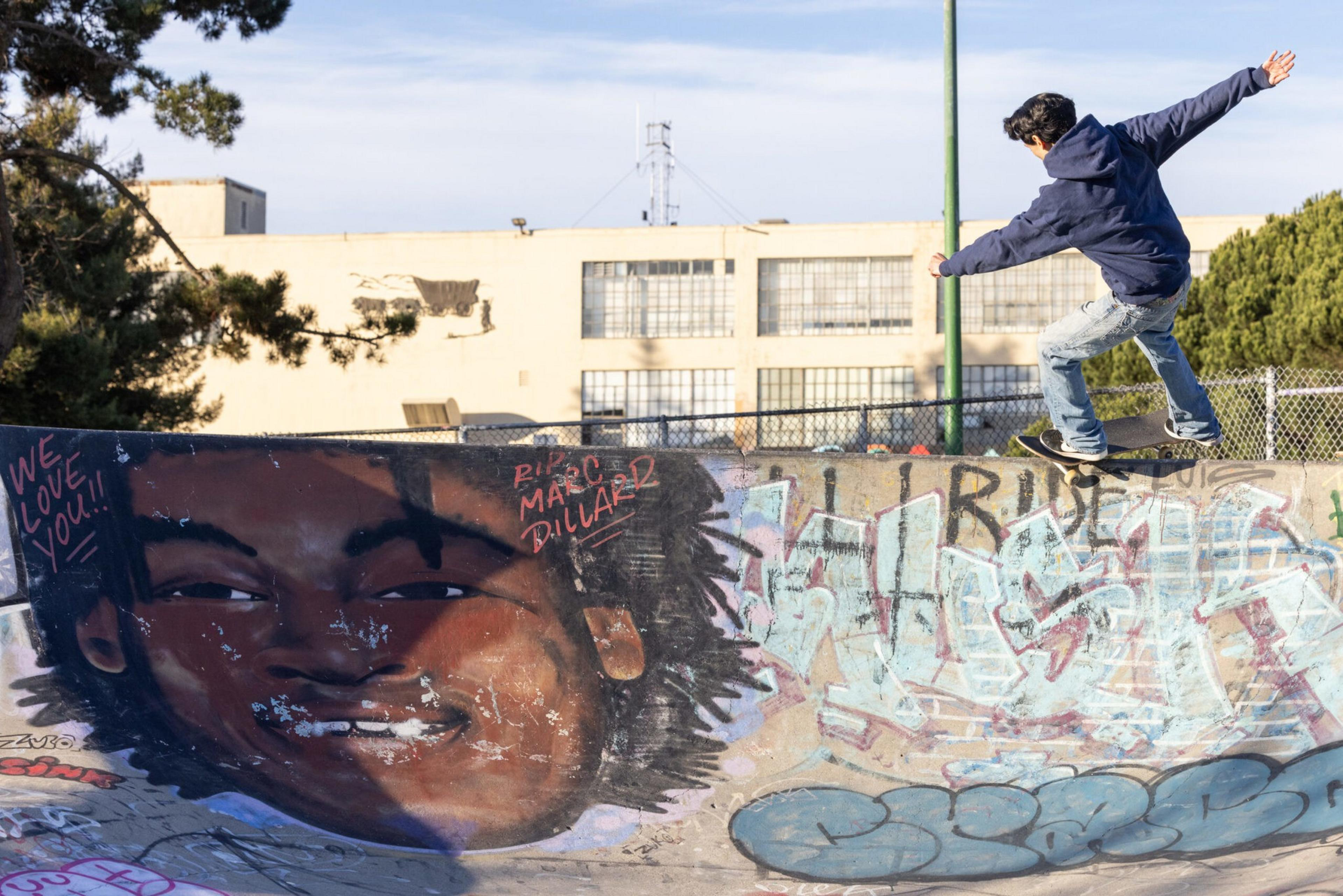 A skateboarder in a blue hoodie and jeans rides along a graffiti-covered concrete skate park featuring a large painted face.