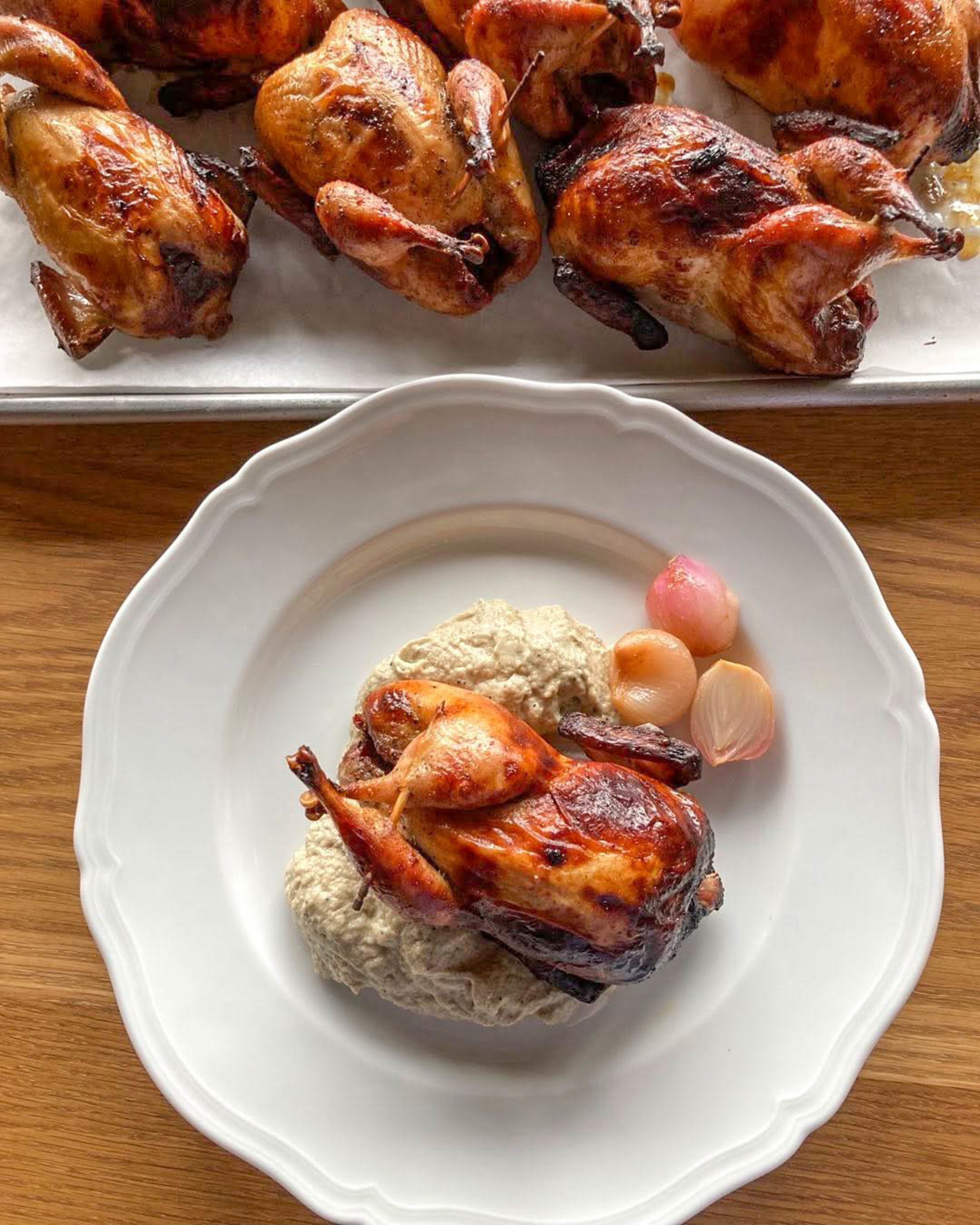 A roasted small bird lies on a creamy mushroom sauce with three peeled shallots on a white plate, with several more birds on a tray in the background.