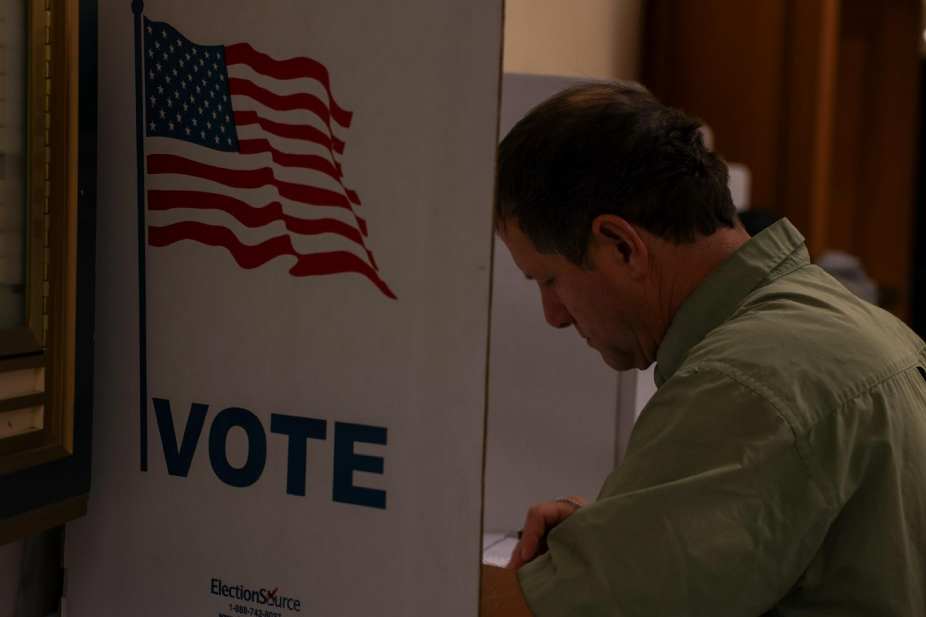 A man is filling out a ballot at a voting booth marked with an American flag and "VOTE."