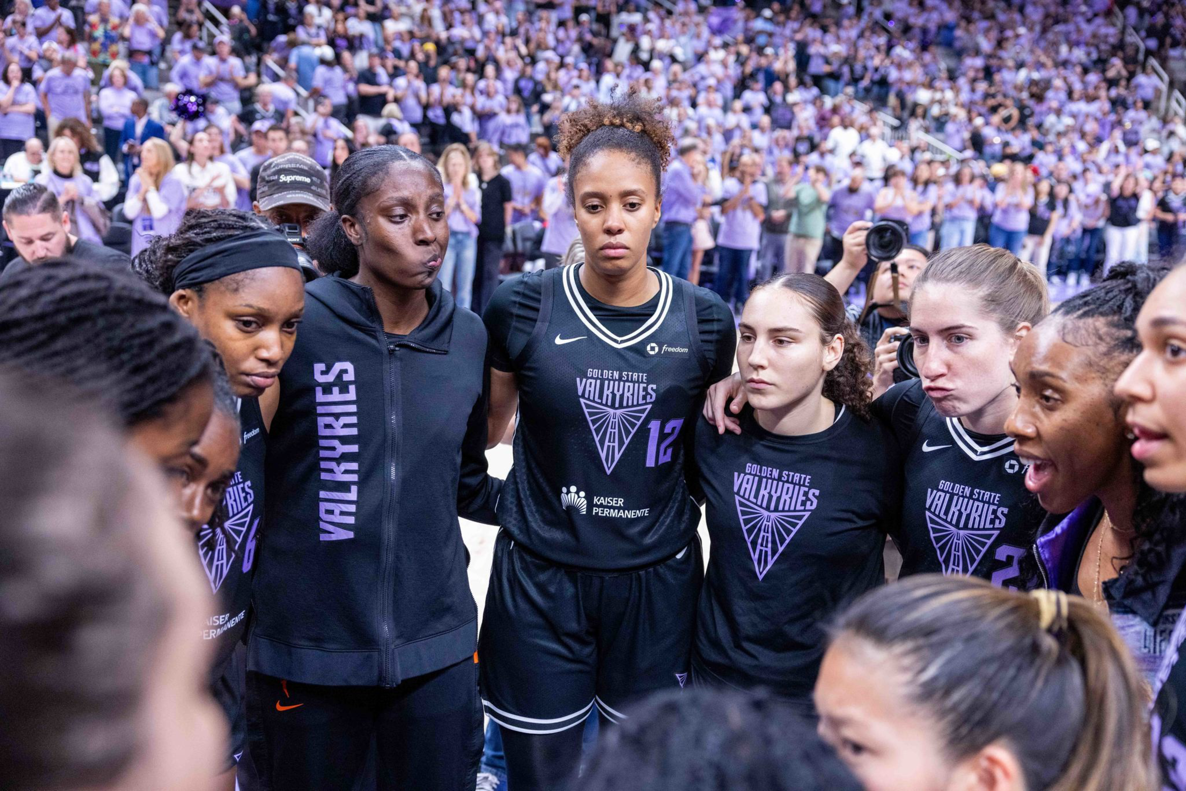 A group of female basketball players wearing Golden State Valkyries uniforms stand closely in a huddle, showing serious and focused expressions.