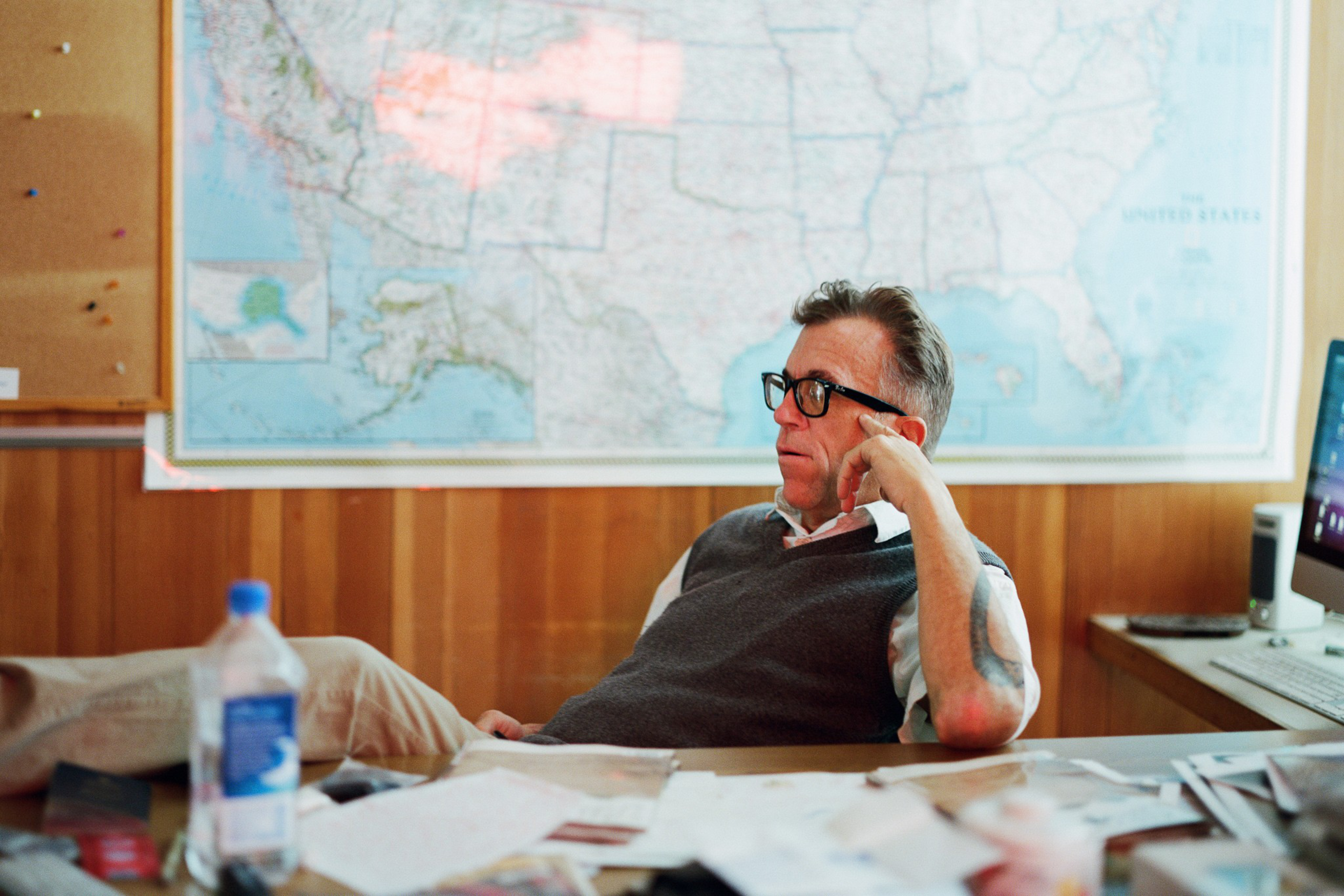 A man wearing glasses and a dark sweater vest sits thoughtfully at a cluttered desk with papers, a water bottle, and a large U.S. map behind him.