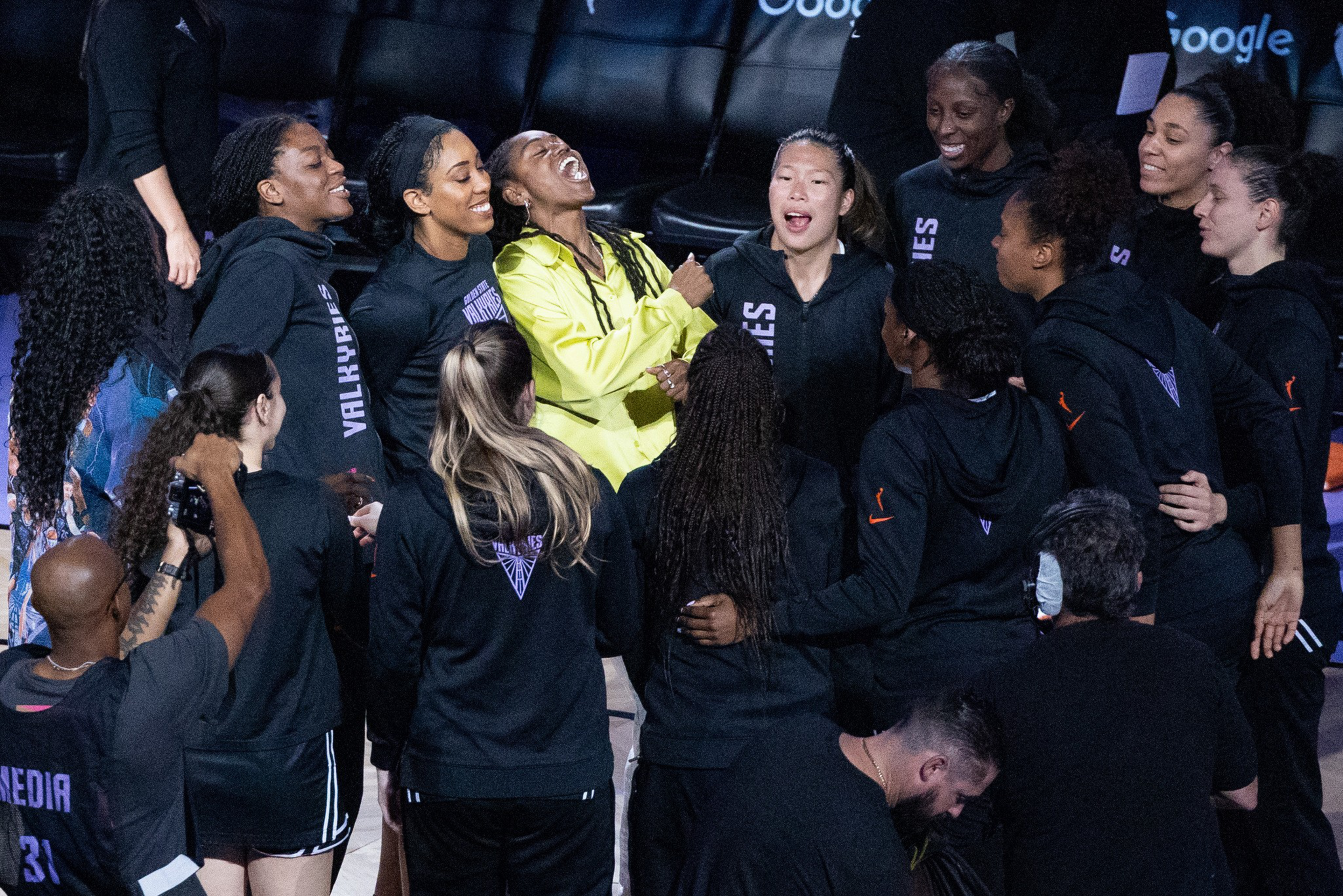 A group of female athletes in black warm-up outfits stand together, smiling and laughing, with one woman in a bright yellow jacket in the center.