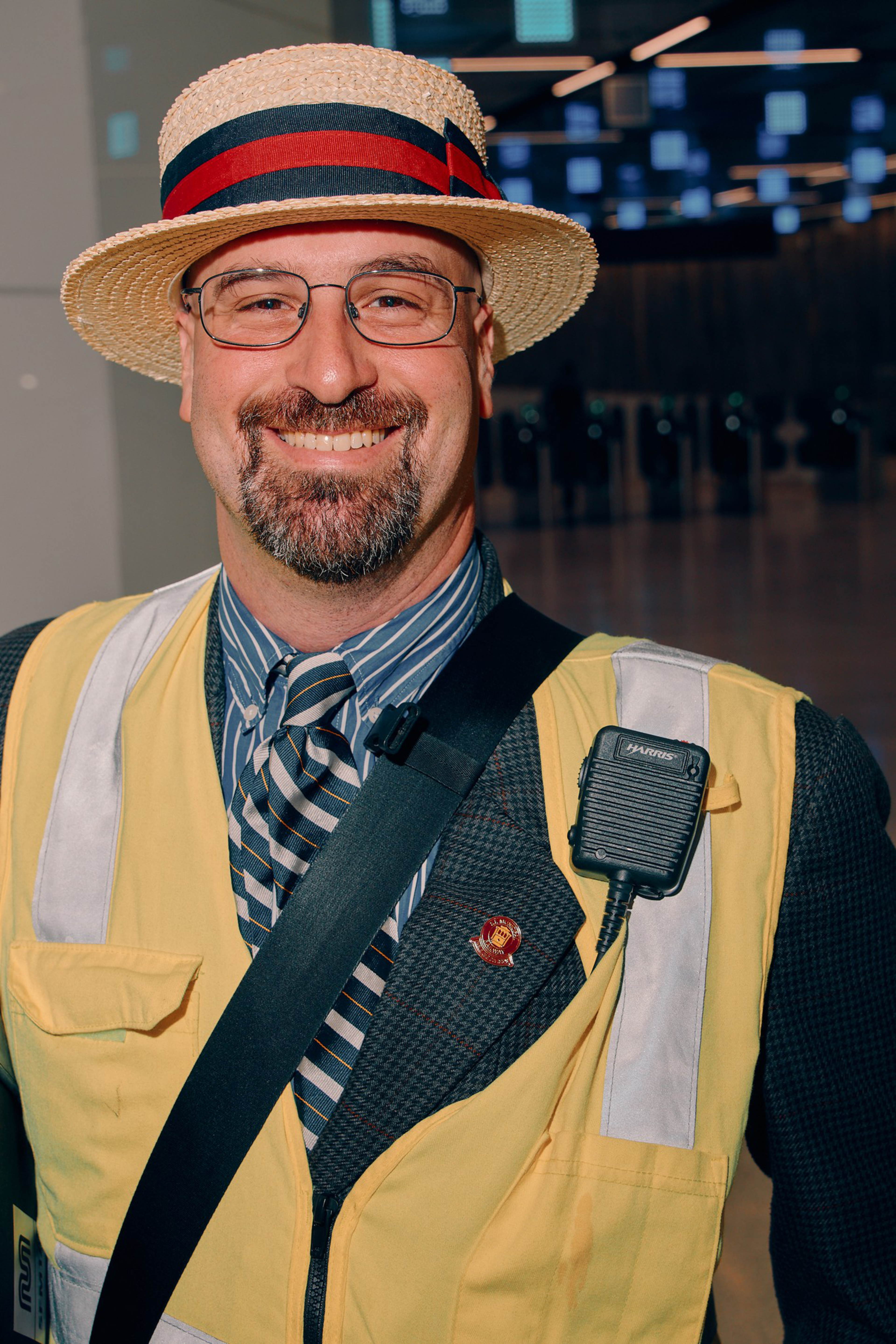 A smiling man wearing glasses, a straw hat with a red and black band, a striped tie, a dark suit, and a yellow safety vest with a walkie-talkie.