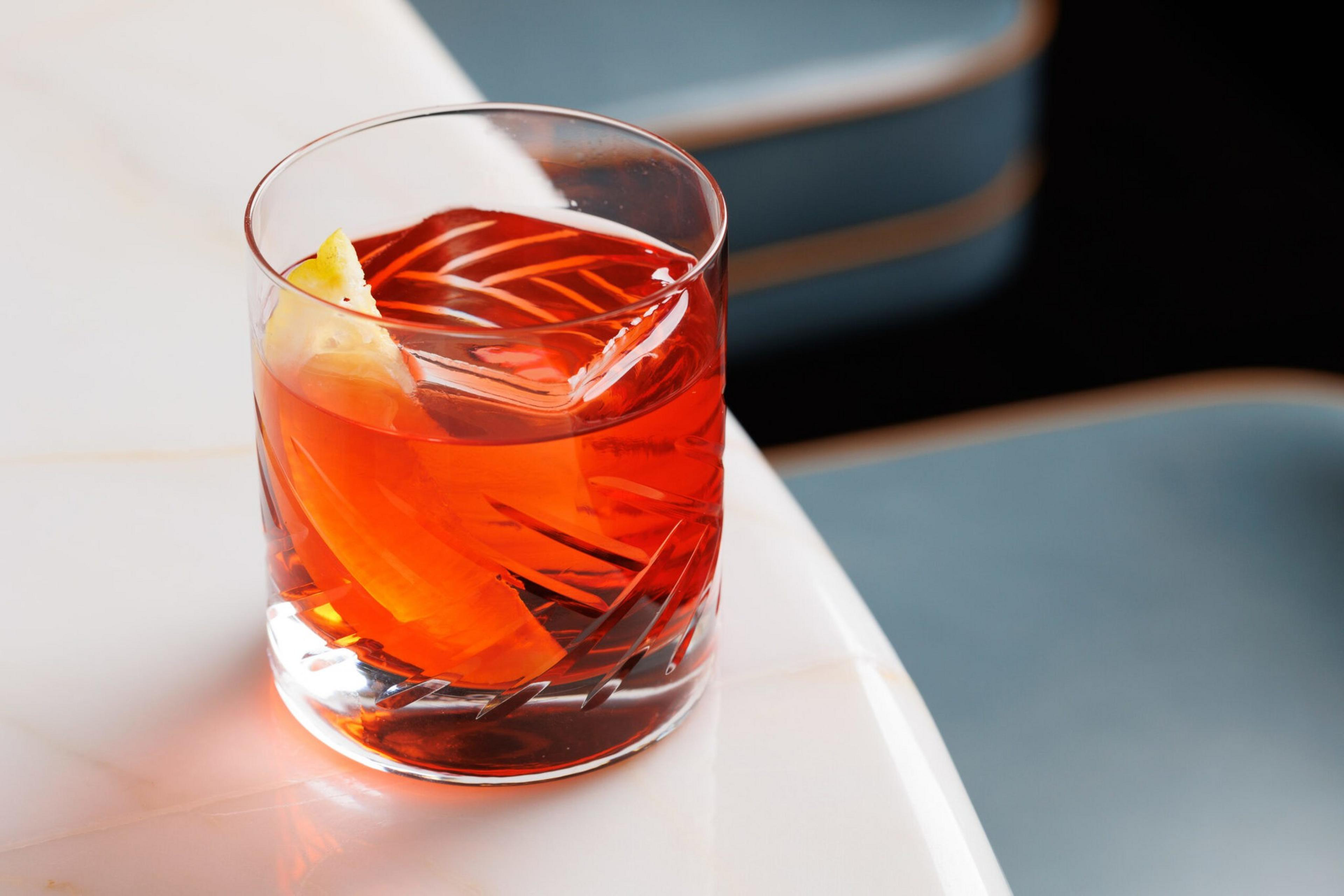 A glass with orange-red liquid, a large ice cube, and a lemon peel sits on a white marble table with blue chairs in the background.