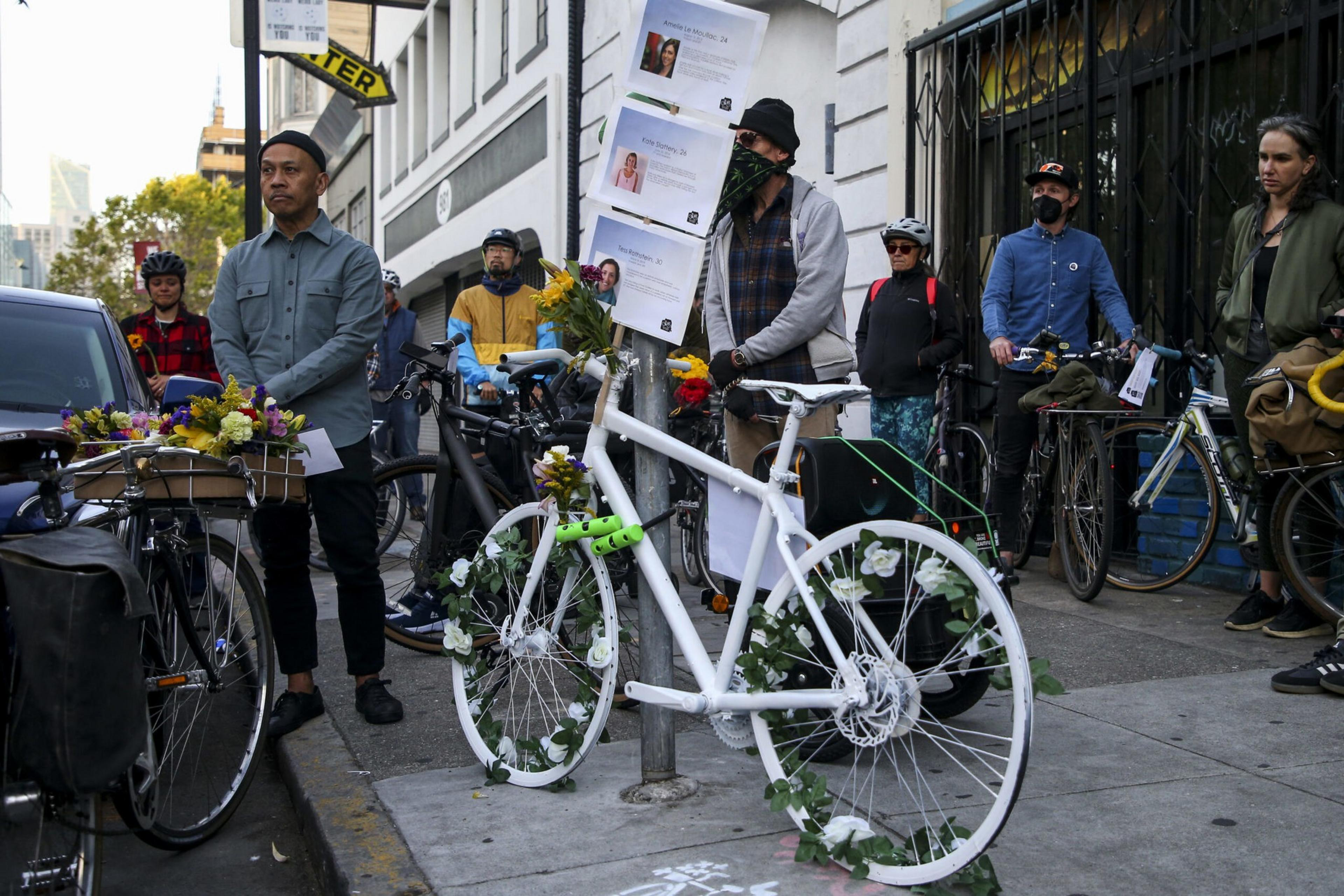 A group of people stand solemnly near a white-painted bicycle adorned with flowers, symbolizing a memorial on a city sidewalk.