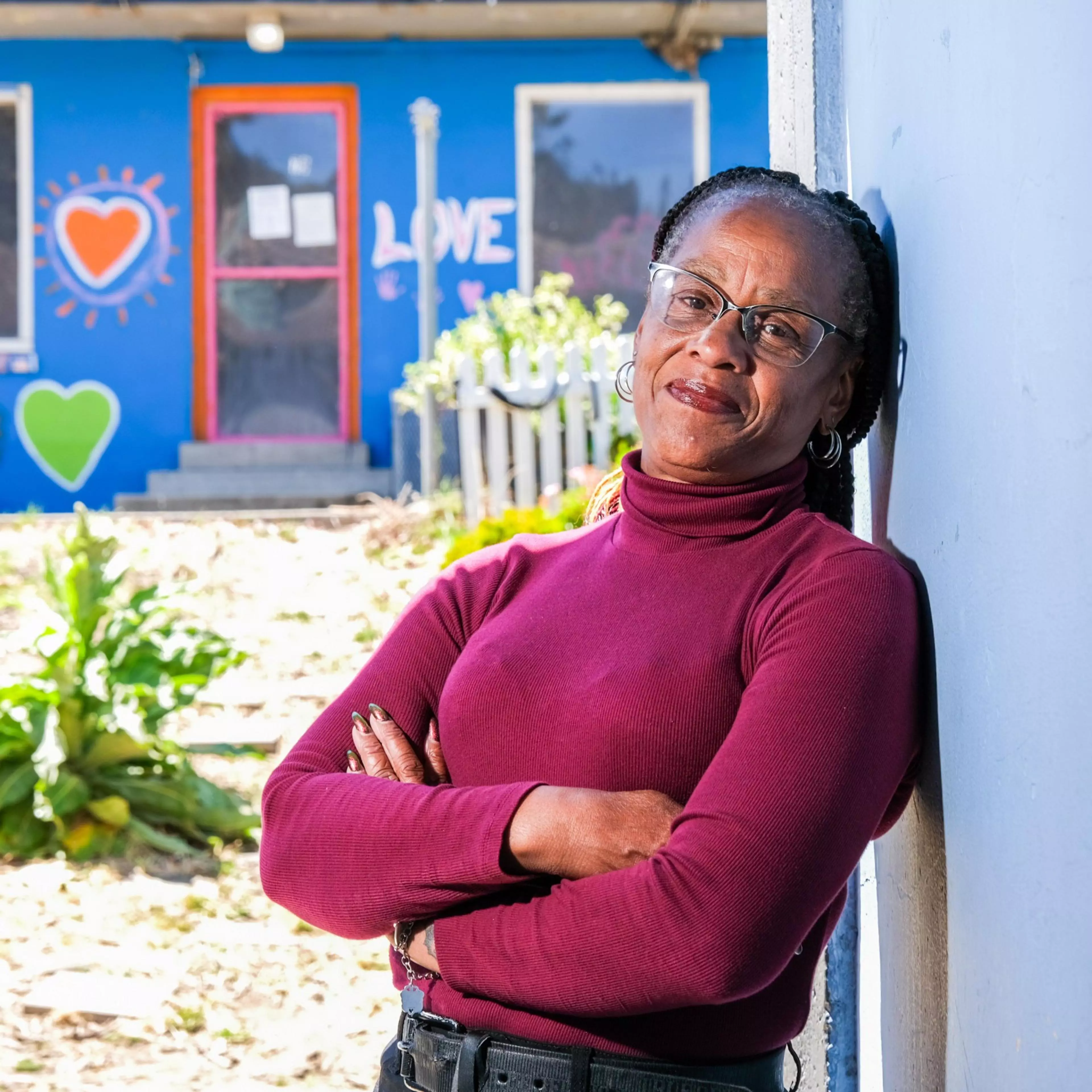 A woman in a burgundy turtleneck, wearing glasses, leans against a wall with her arms crossed. The background shows a blue building with colorful heart and love decorations.
