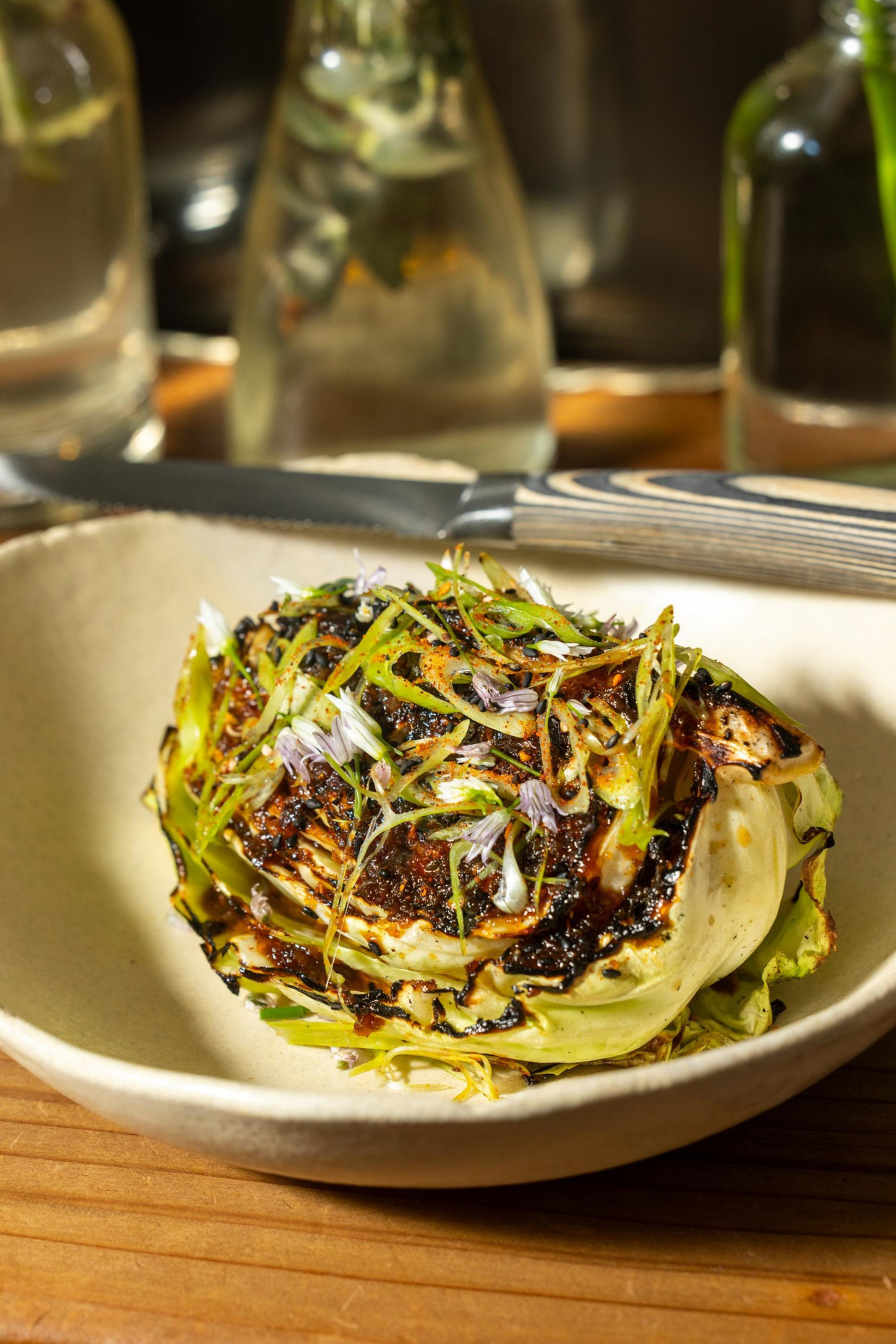 A charred cabbage wedge garnished with thinly sliced green herbs and small purple flowers rests in a beige bowl on a wooden table.