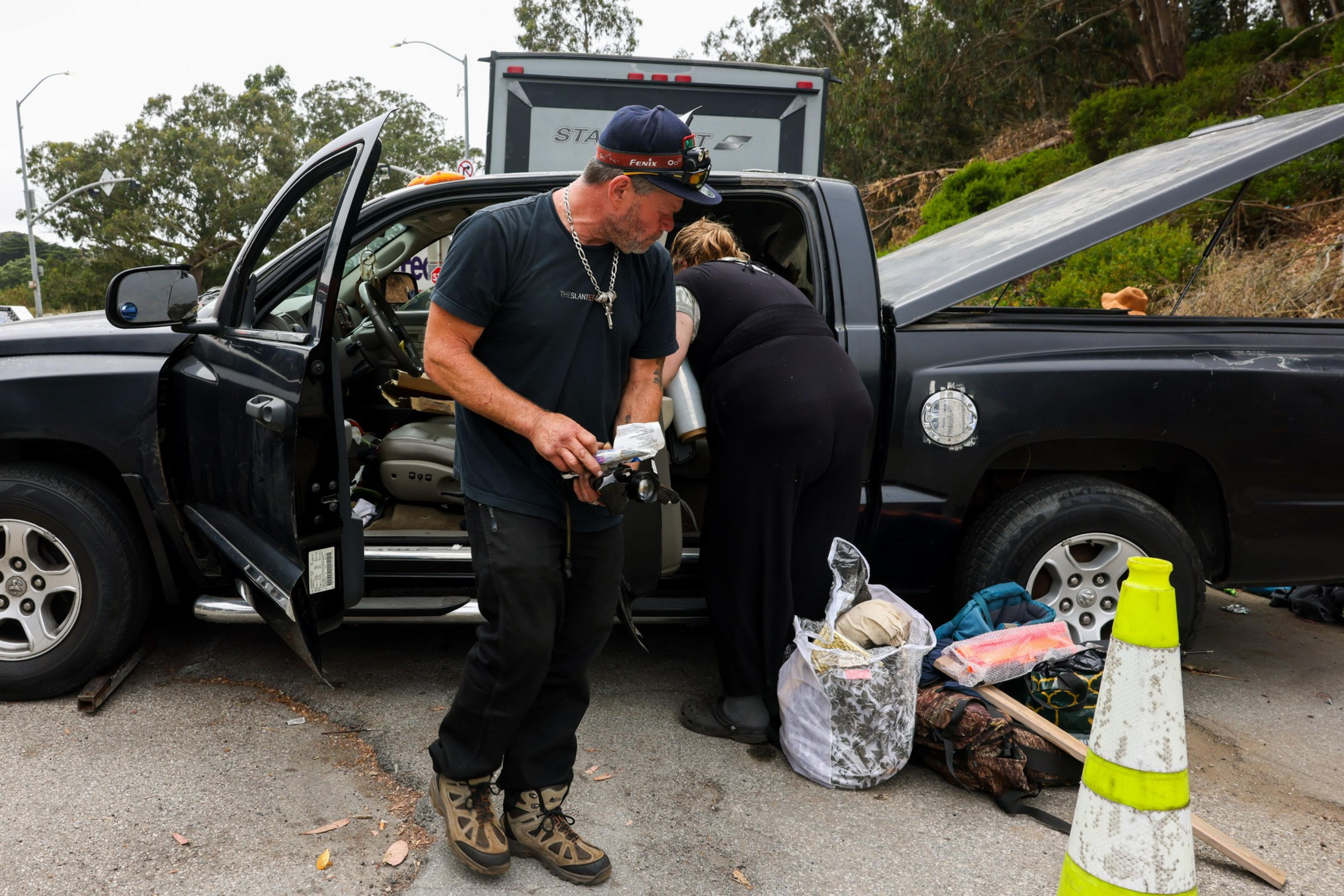A man and woman organize items from an open truck parked on a roadside. Various bags and a yellow traffic cone are on the ground nearby.