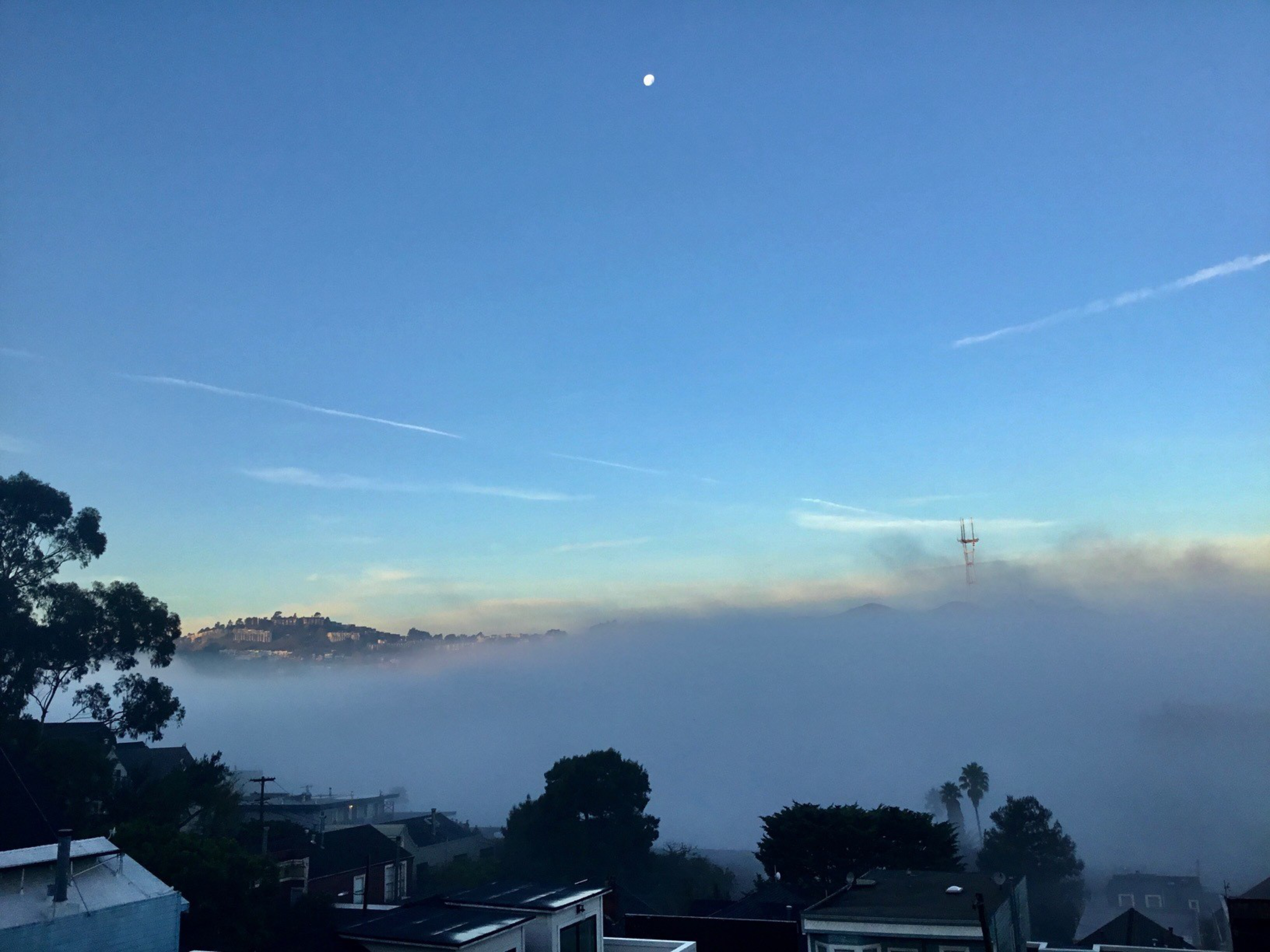 A serene scene shows a moonlit sky with light clouds over a foggy landscape, silhouetted trees, rooftops, and a distant hill partially covered by mist.