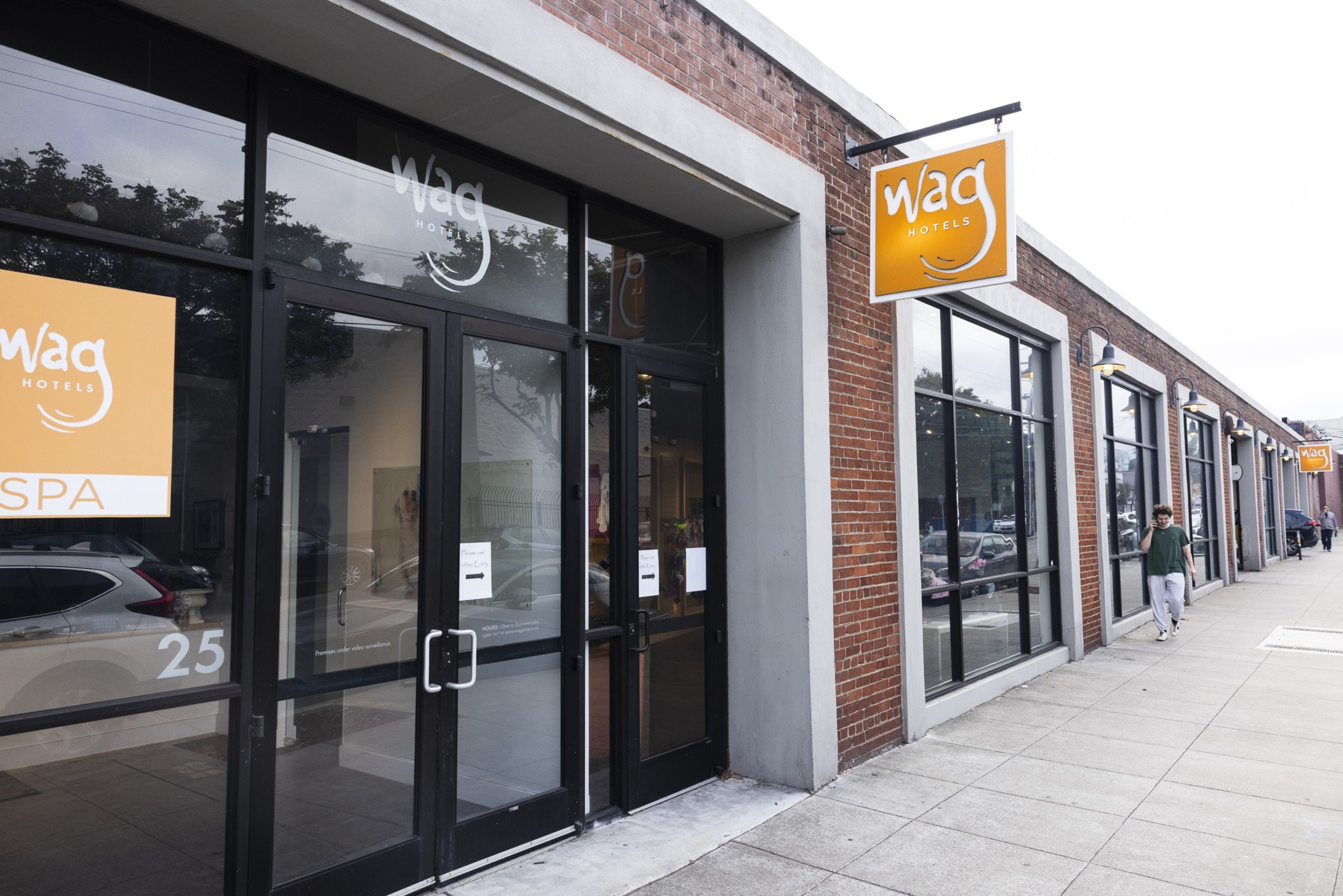 A brick building with large windows and multiple orange "wag hotels" signs lines a sidewalk where a person in a green shirt and white pants is walking.