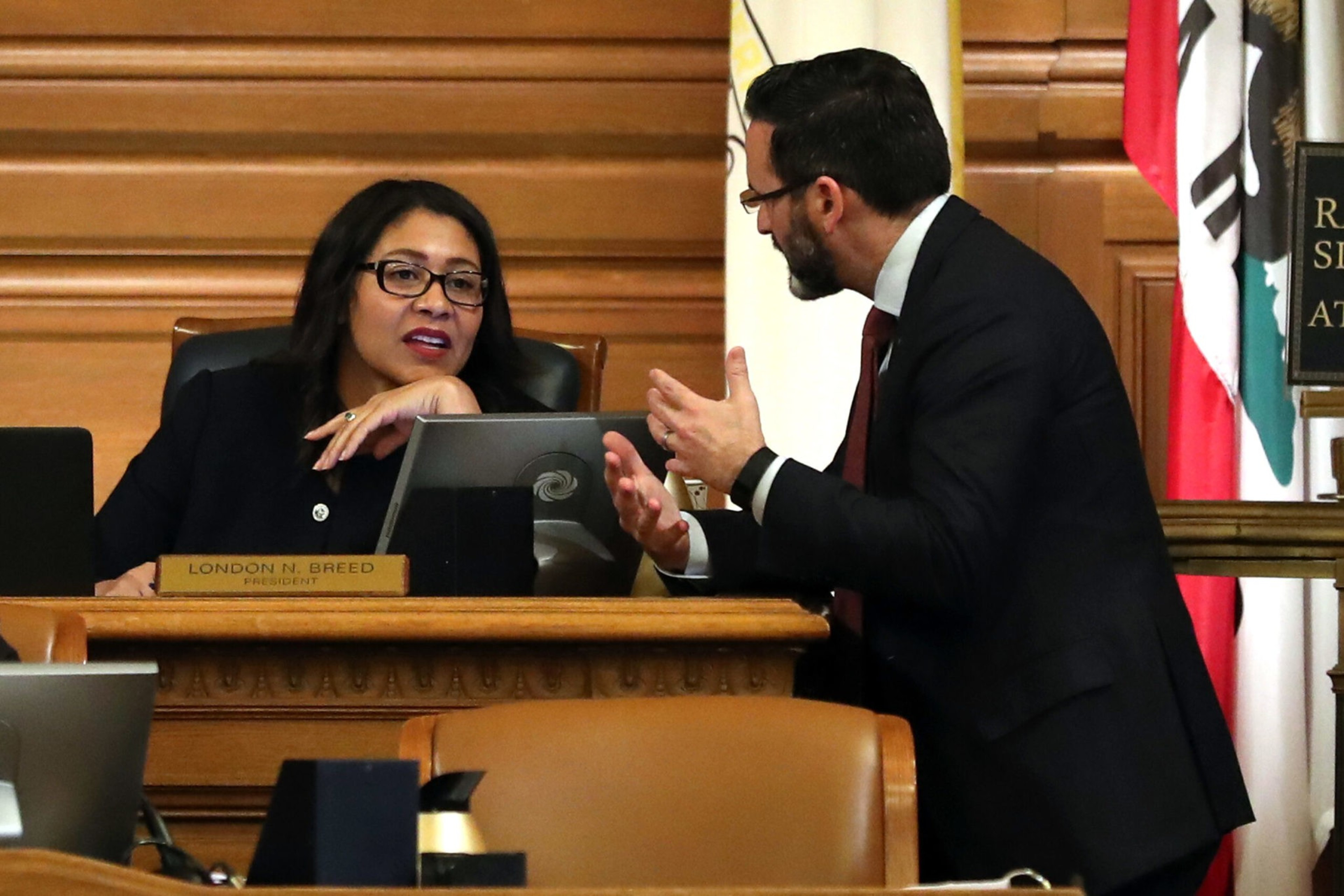 A woman, seated behind a desk with a nameplate reading "London N. Breed President," listens as a man in a suit gestures while speaking to her.