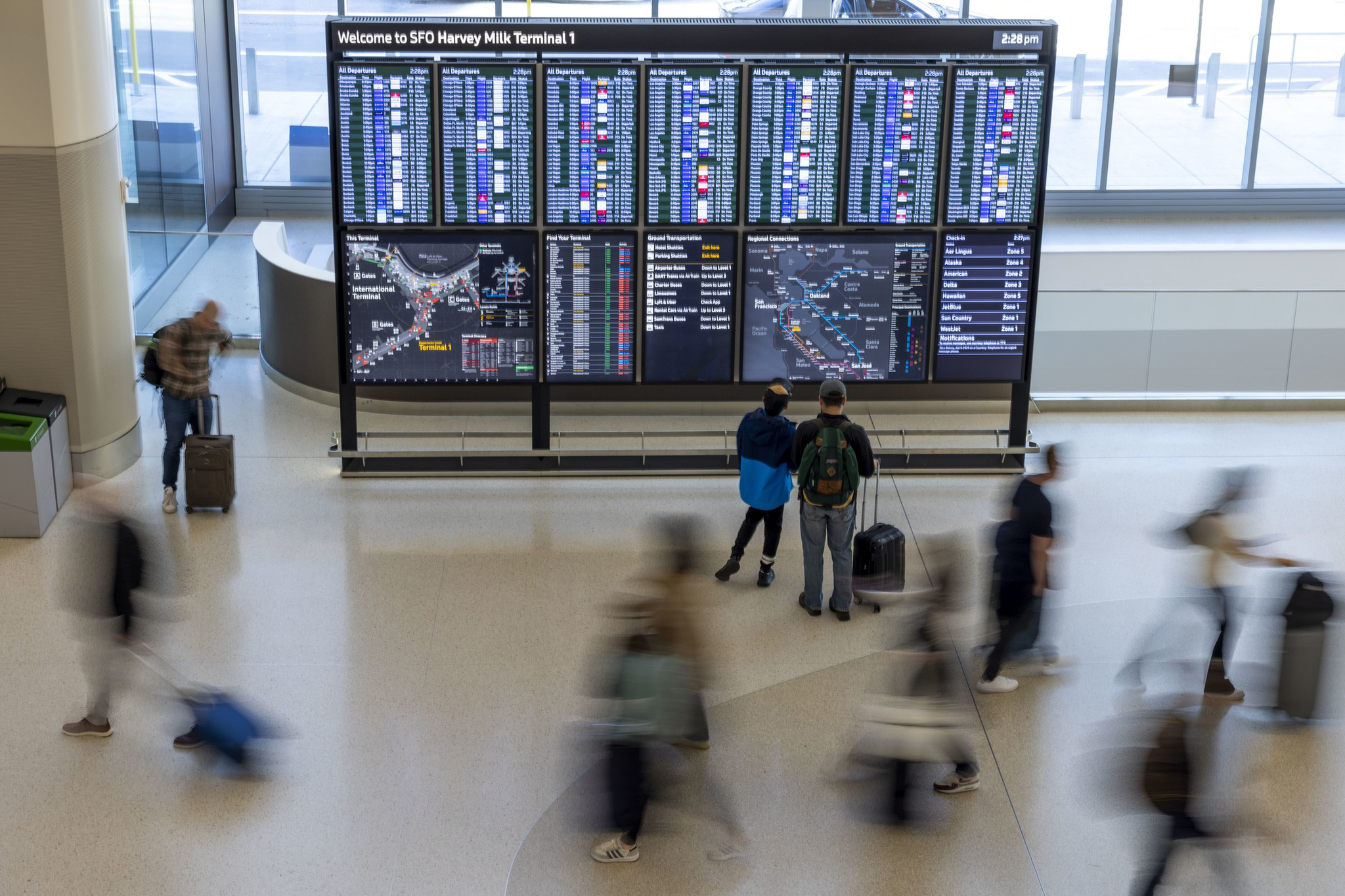 A large flight information board at SFO Terminal 1 displays arrivals and departures as travelers with suitcases move around, some blurred in motion.