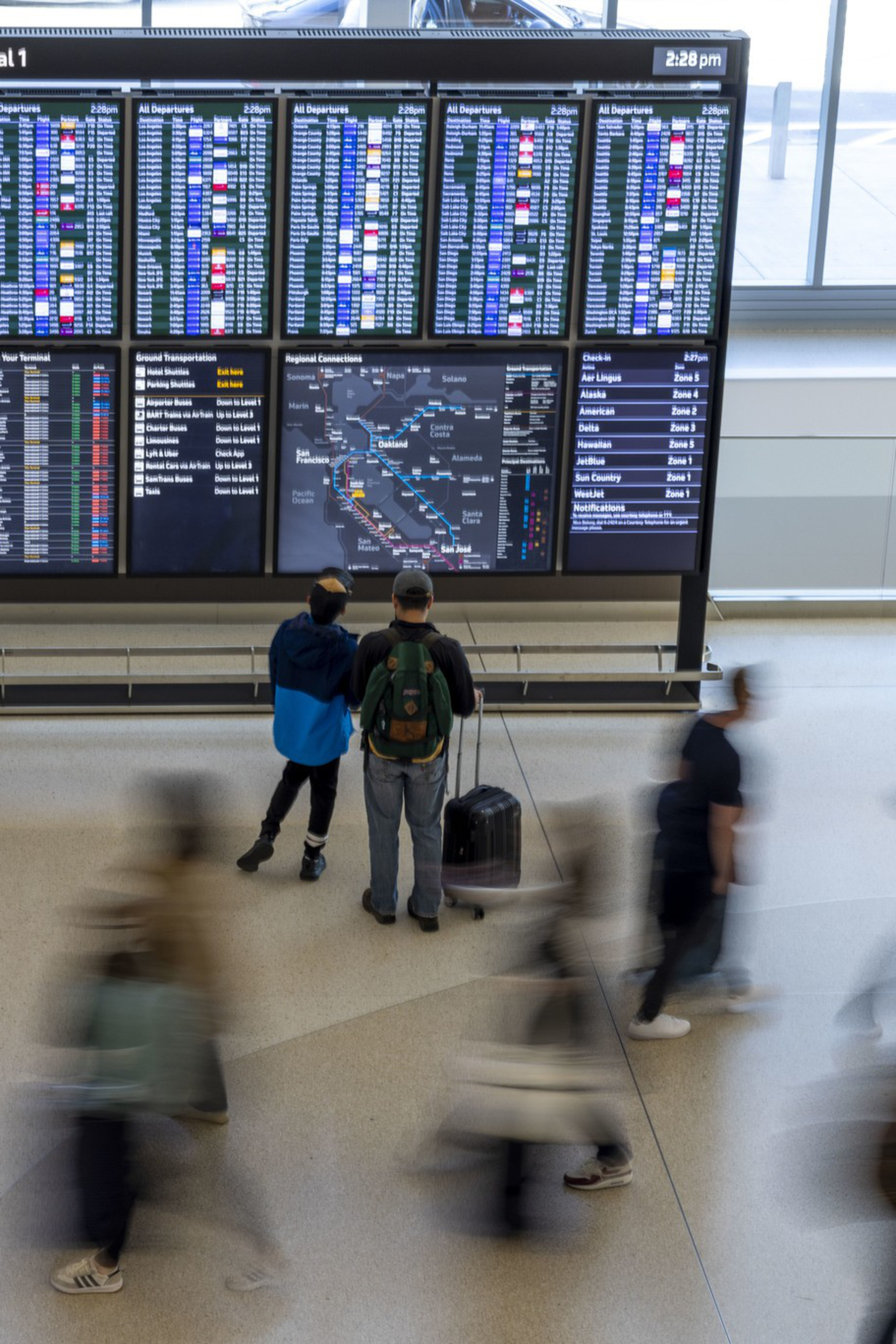 A large flight information board at SFO Terminal 1 displays arrivals and departures as travelers with suitcases move around, some blurred in motion.