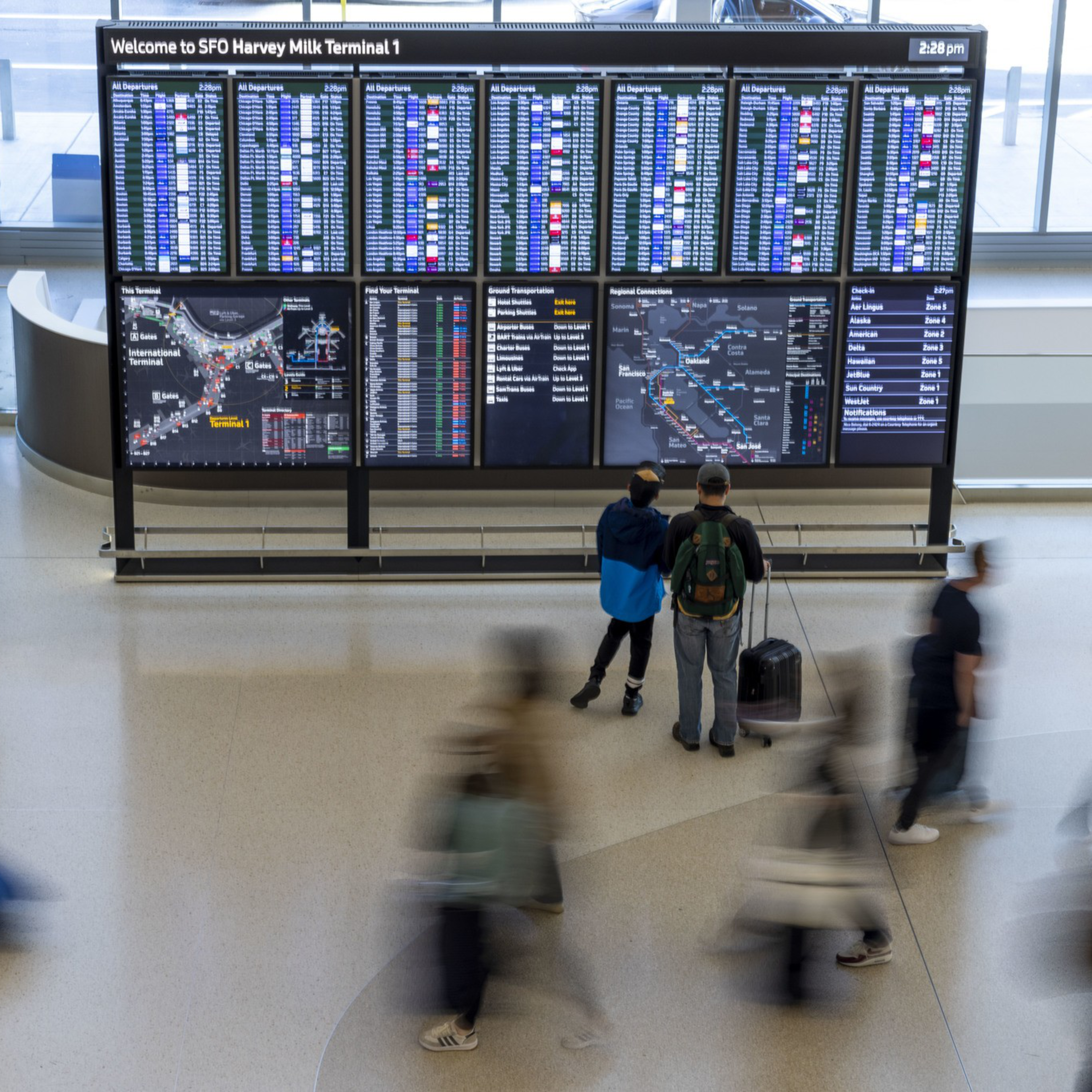 A large flight information board at SFO Terminal 1 displays arrivals and departures as travelers with suitcases move around, some blurred in motion.