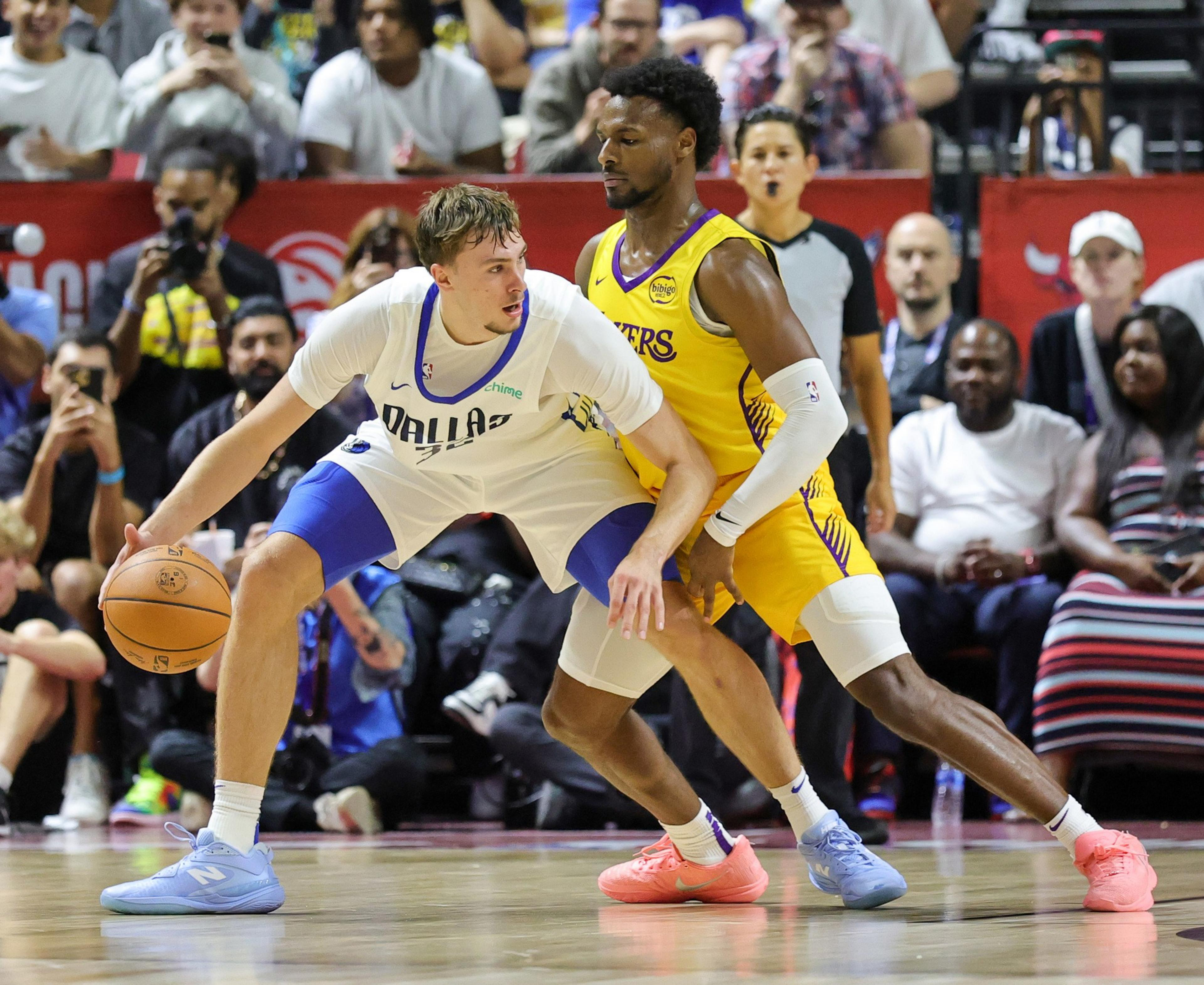 Two basketball players are on the court, with one in a Dallas uniform dribbling the ball and the other in a Lakers uniform defending. Spectators watch closely.