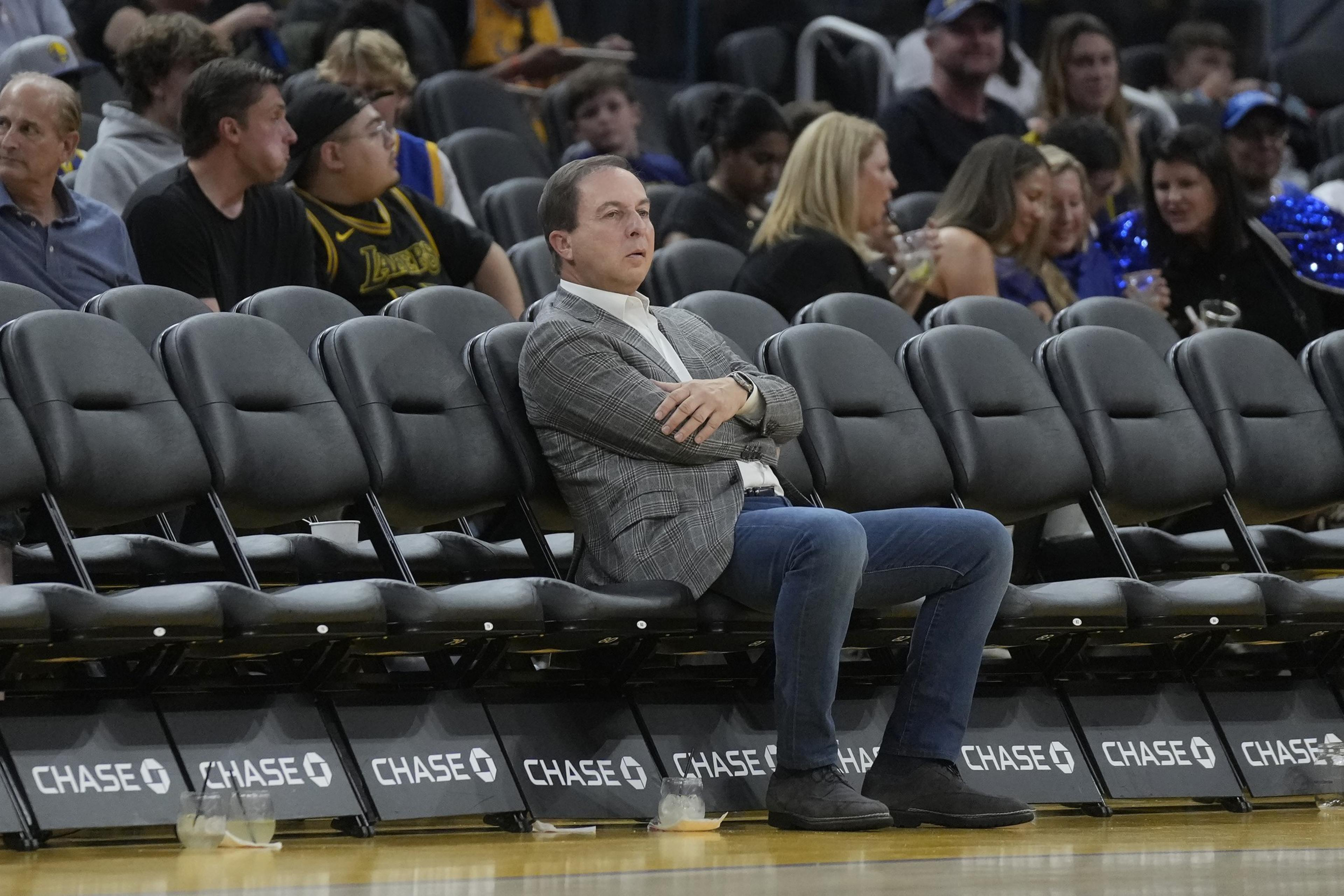 A man in a gray suit and crossed arms sits pensively on an empty row of black seats at a sports event, with a lively crowd behind him.