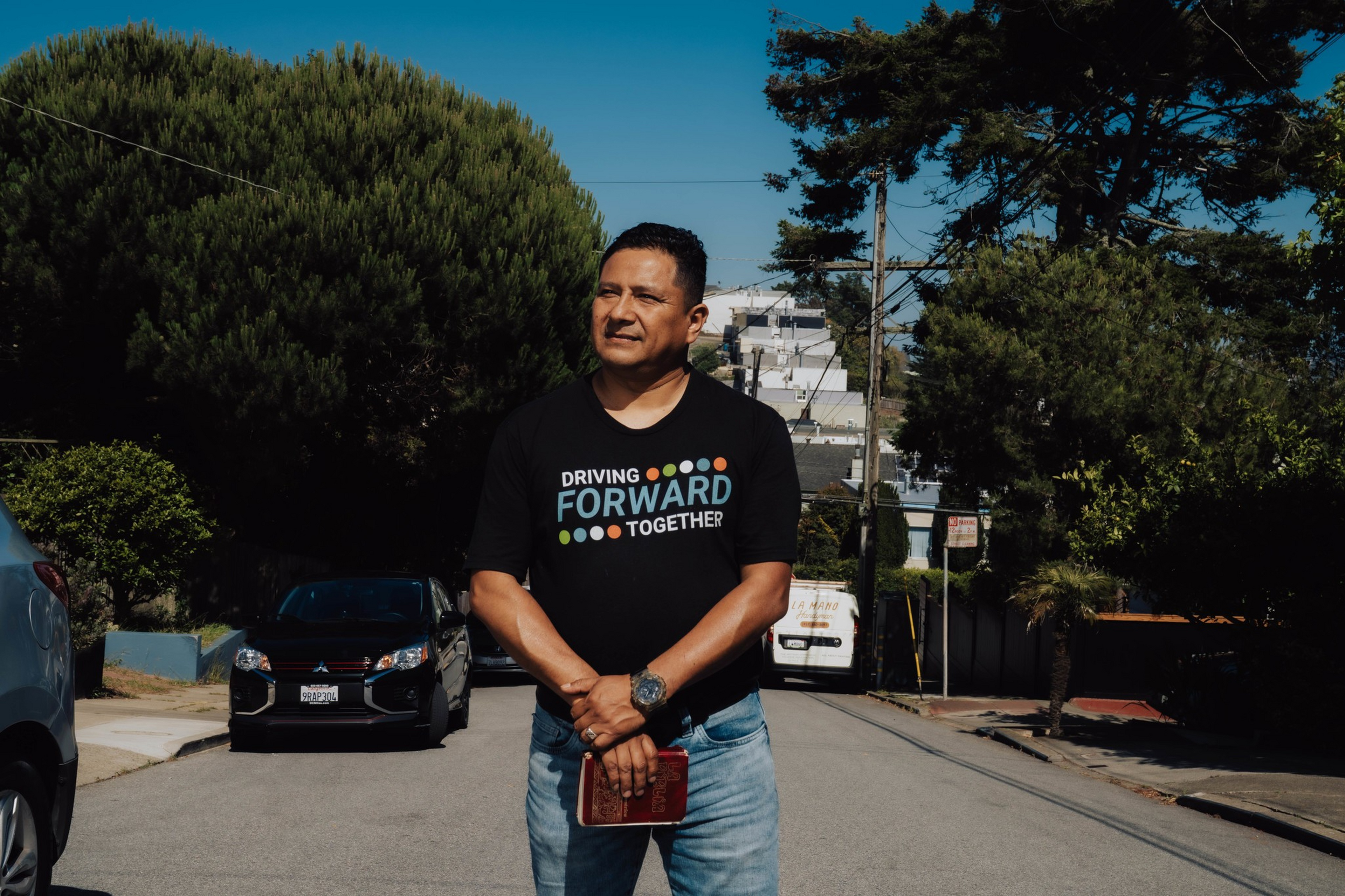 A man stands on a quiet street with trees and houses in the background, wearing a black T-shirt that says "Driving Forward Together."