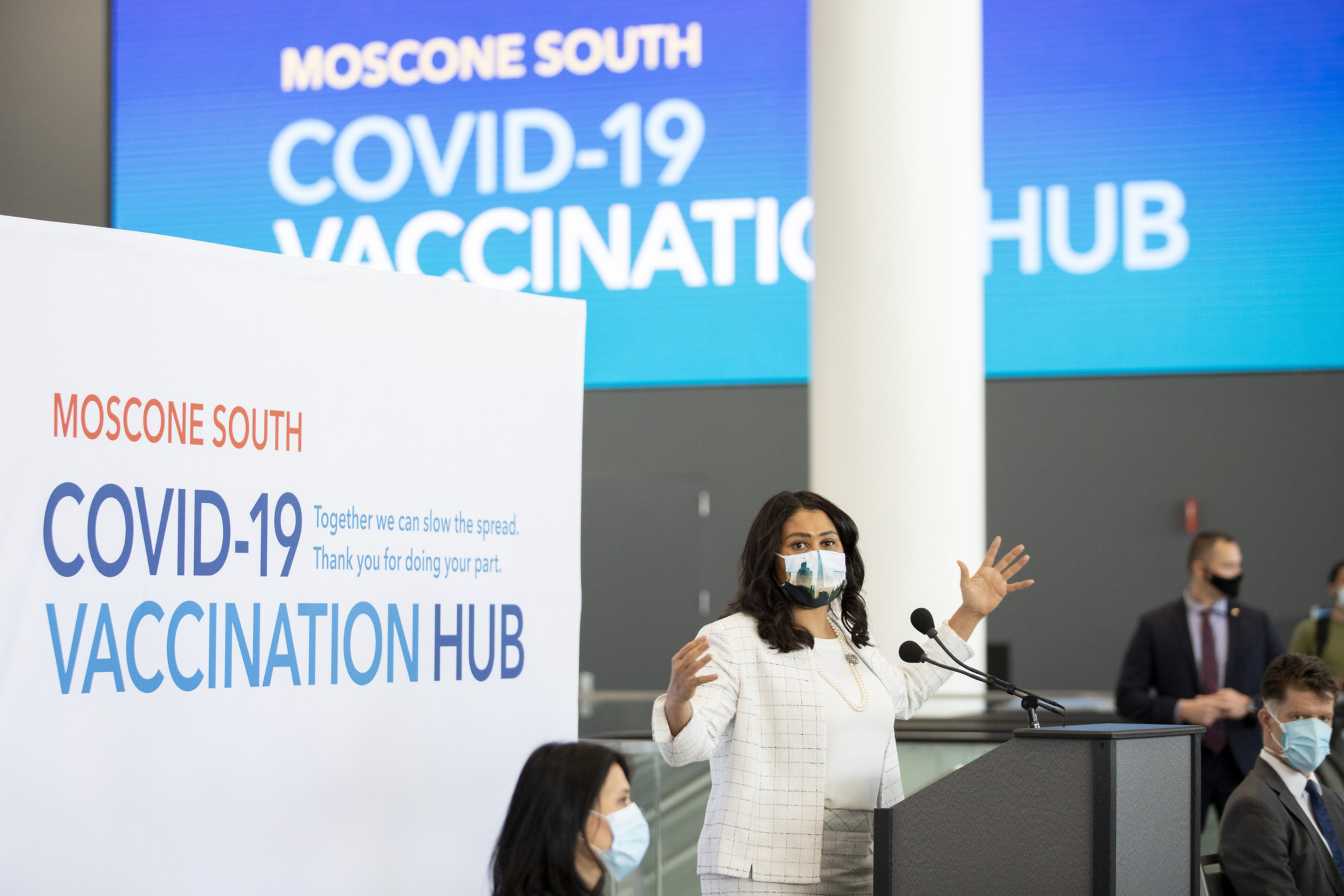 A woman speaks at a podium wearing a mask in a COVID-19 vaccination hub, with a large sign and backdrop displaying &quot;MOSCONE SOUTH COVID-19 VACCINATION HUB.&quot;