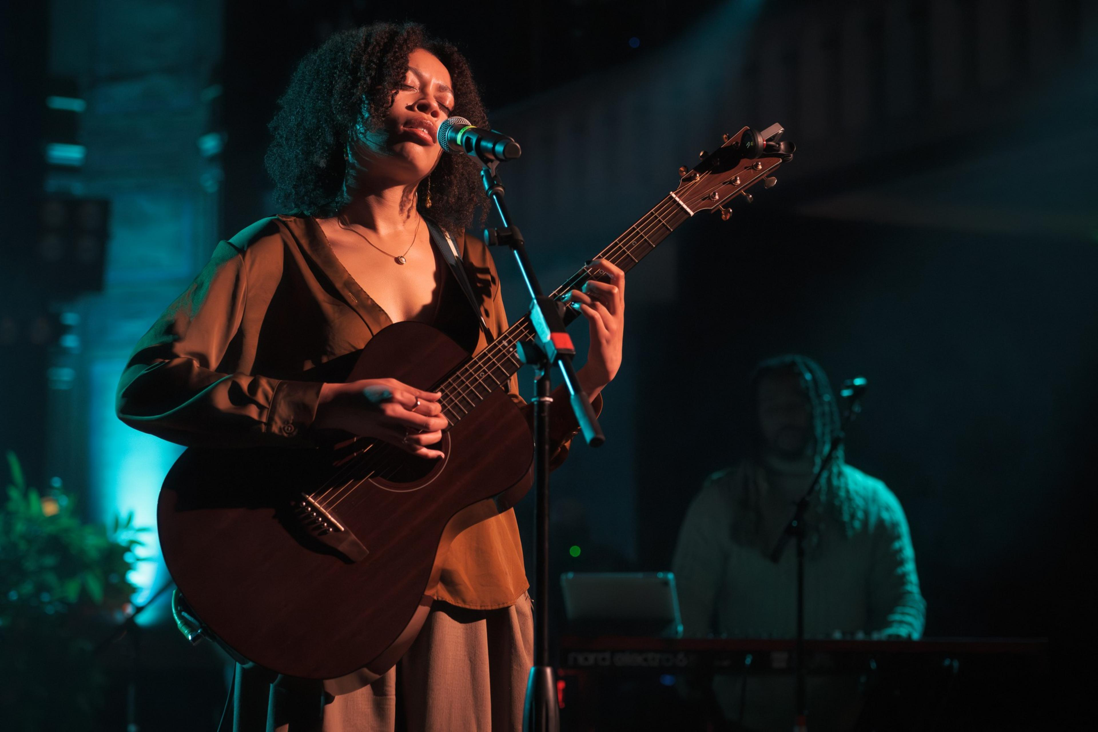 A woman with curly hair plays an acoustic guitar and sings into a microphone. She's illuminated by colorful lighting, with a keyboardist in the background.