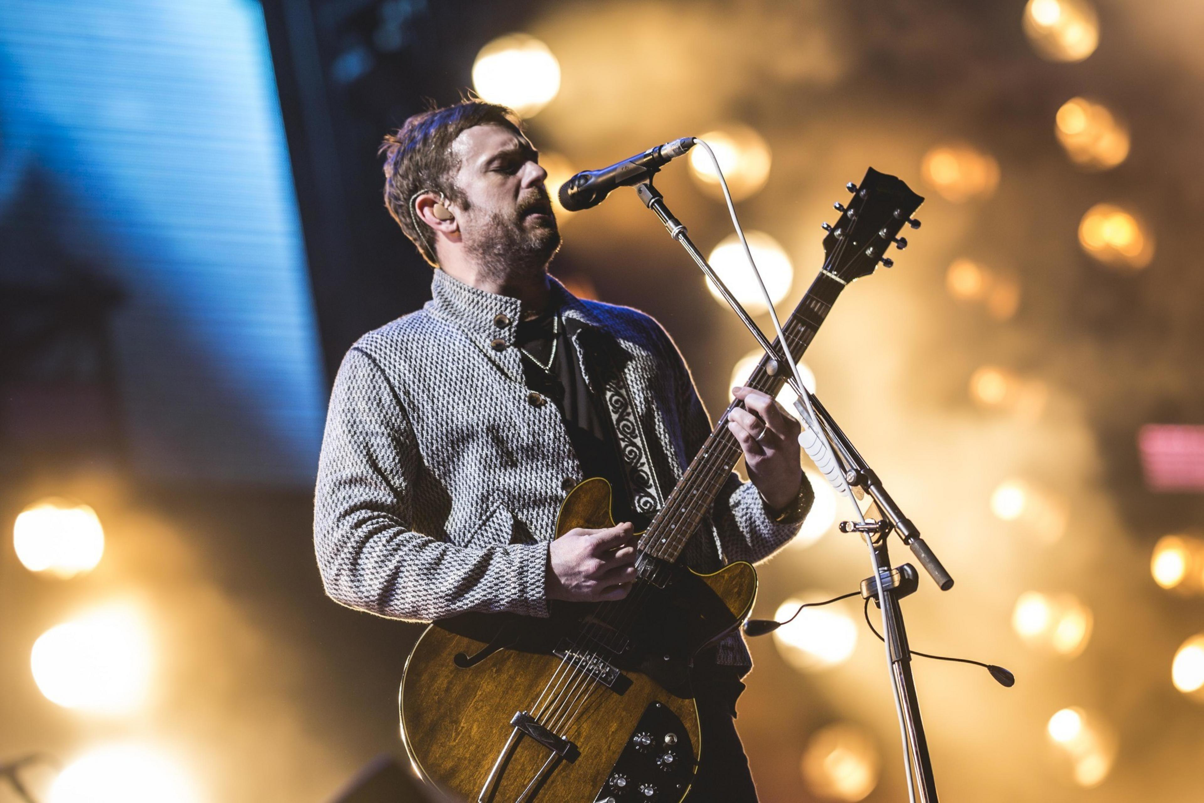 A man is playing an electric guitar and singing into a microphone on stage. He's wearing a patterned jacket, with bright stage lights glowing behind him.