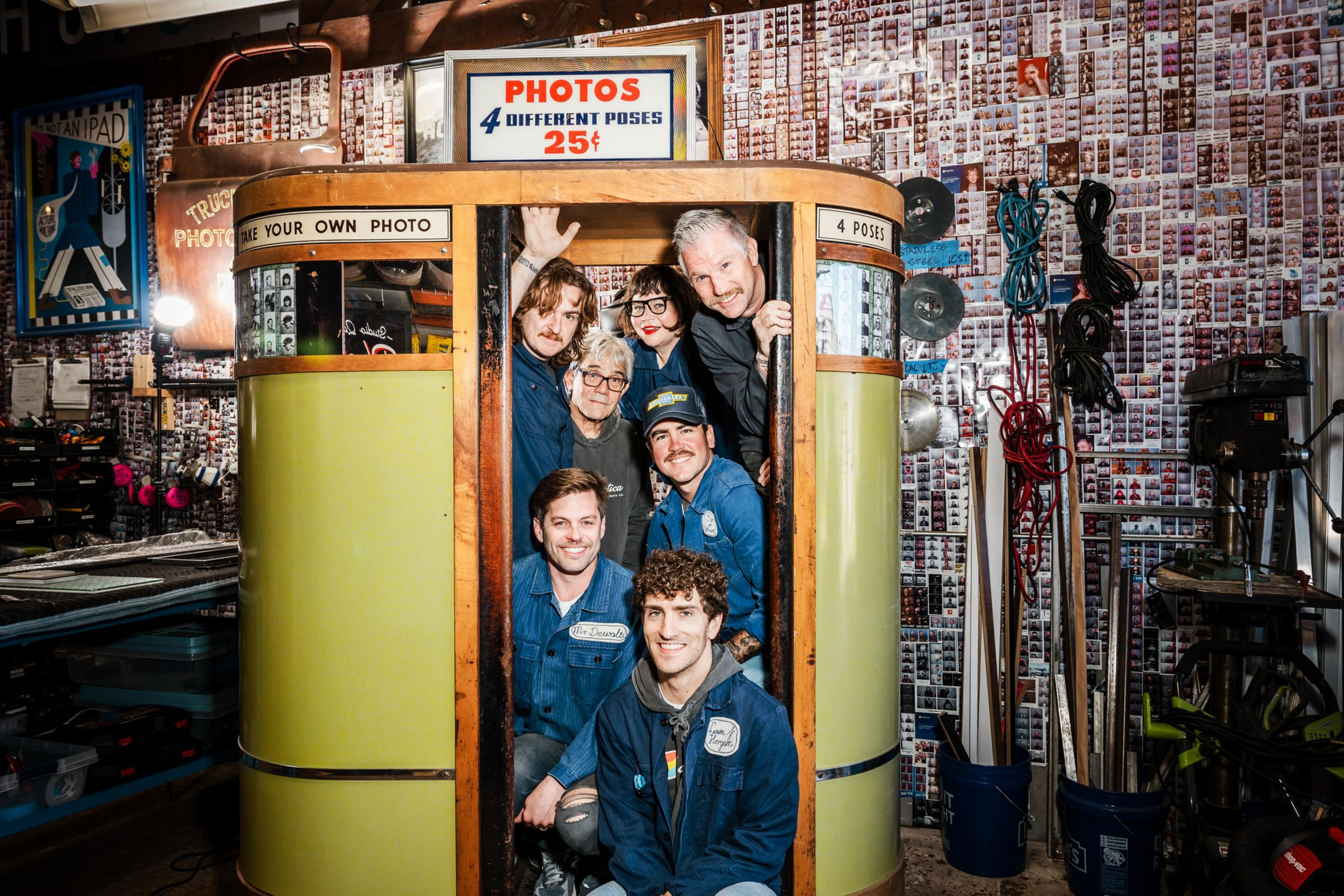 A vintage photo booth in a cluttered workshop displays a sign offering 4 poses for 25 cents. Six smiling people, some in blue overalls, are crowded inside and around the booth.