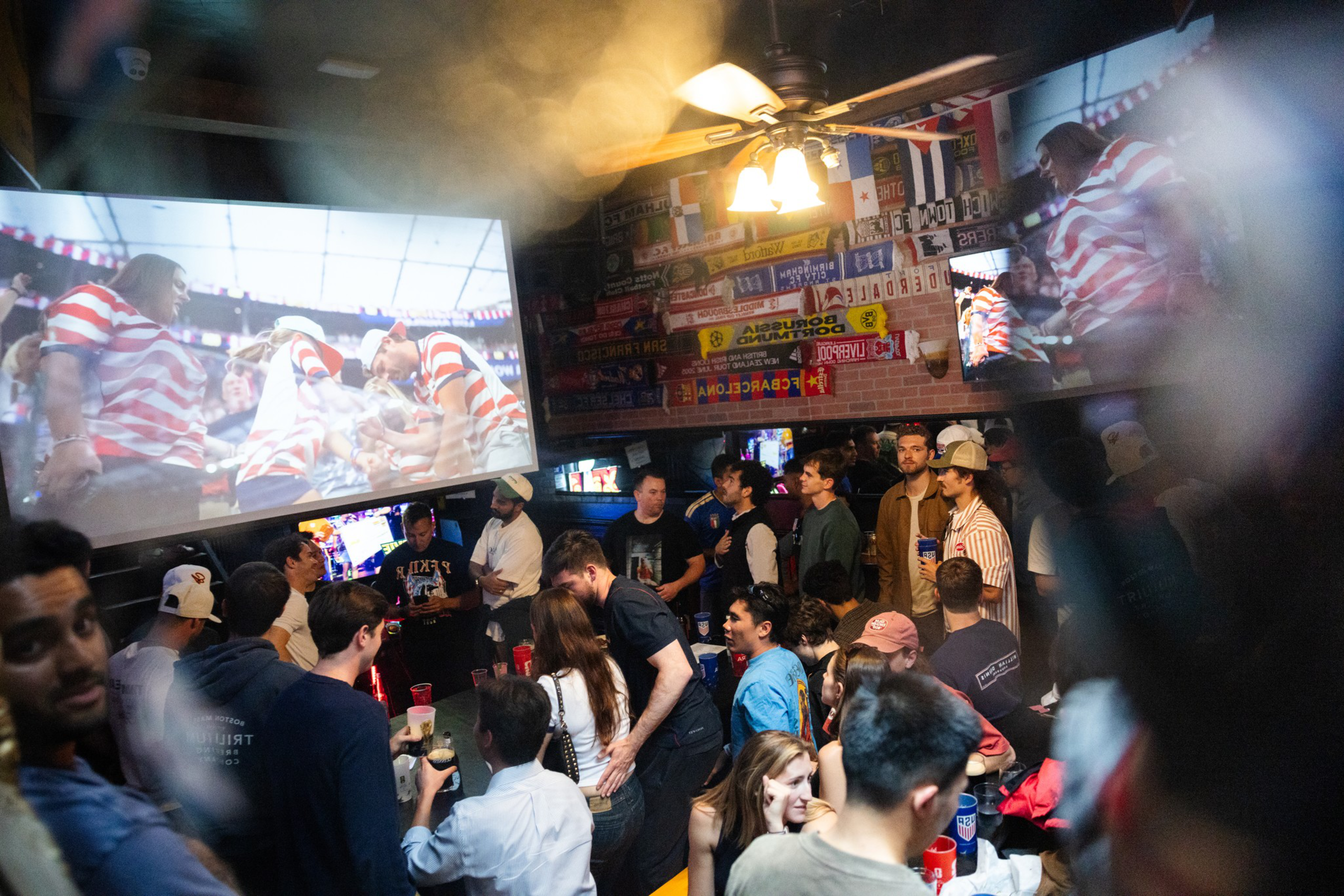 A crowded bar with people watching a sports game on large screens, colorful flags and scarves hanging on the wall, and a ceiling fan overhead.