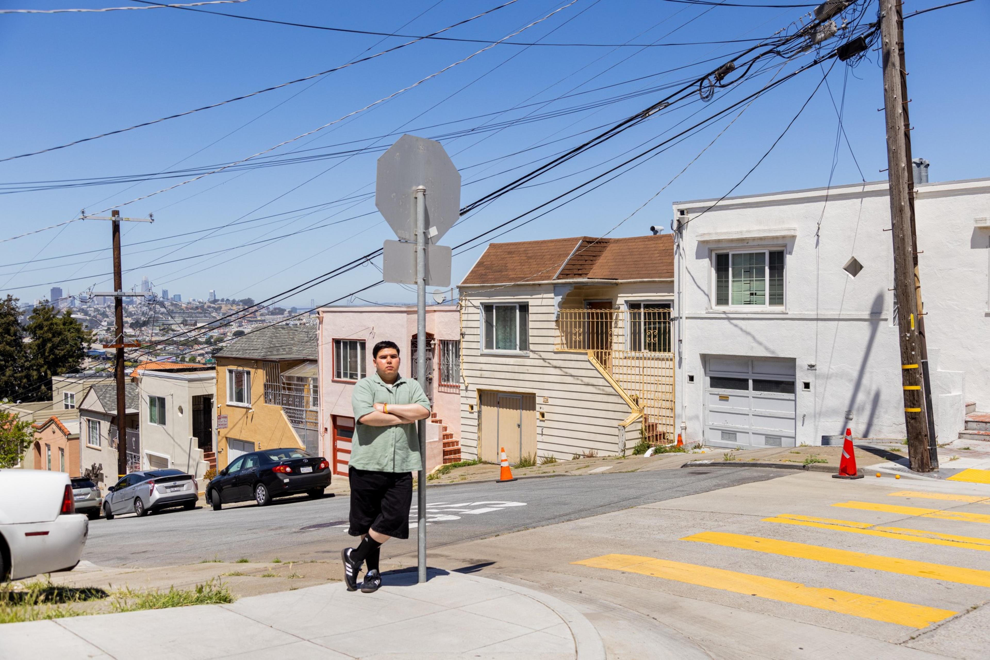A person stands on a sunny street corner with houses and a distant city skyline.
