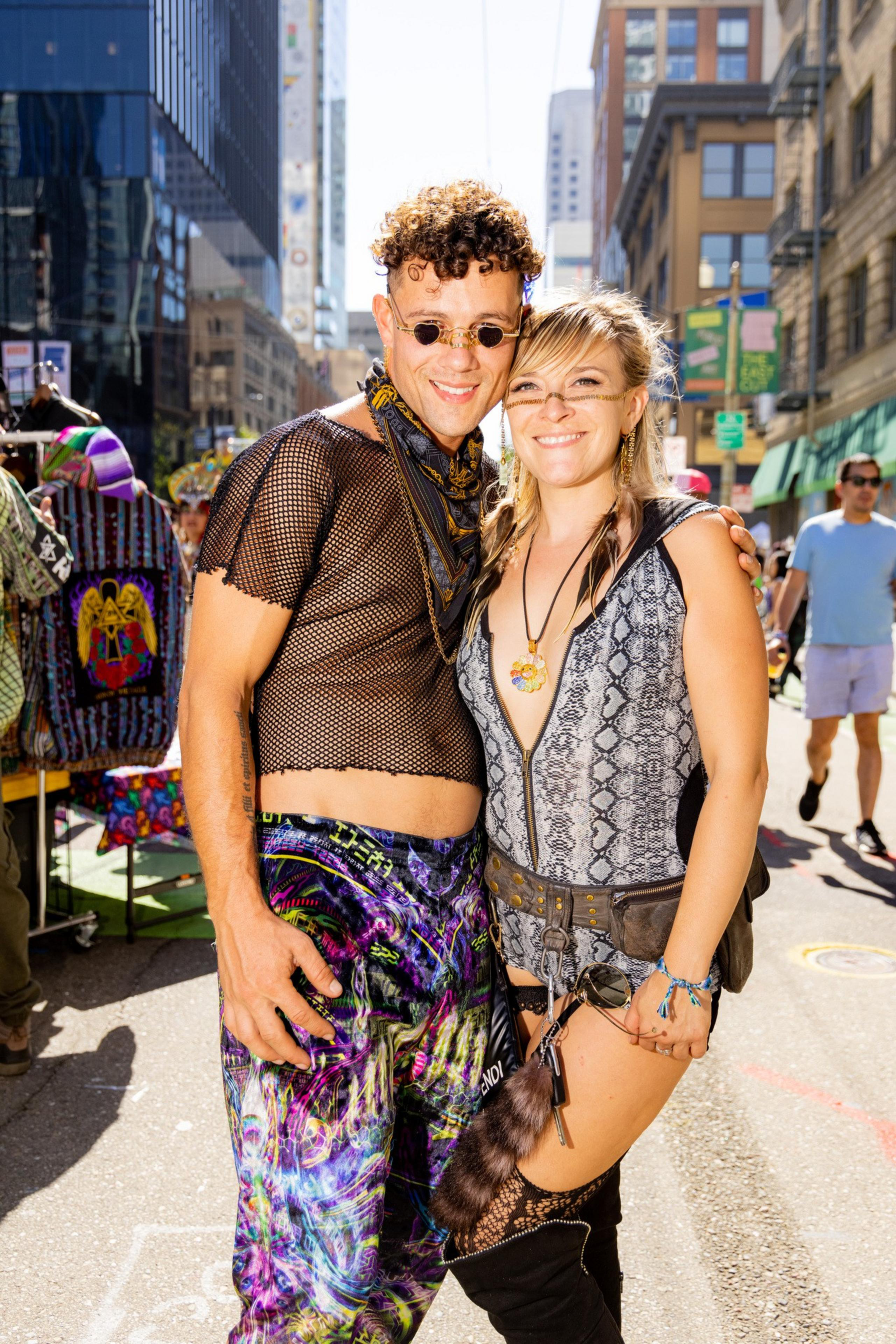 A smiling couple stands side by side in colorful, eclectic outfits on a sunny city street. They appear to be enjoying a festival or street event with people and stalls in the background.