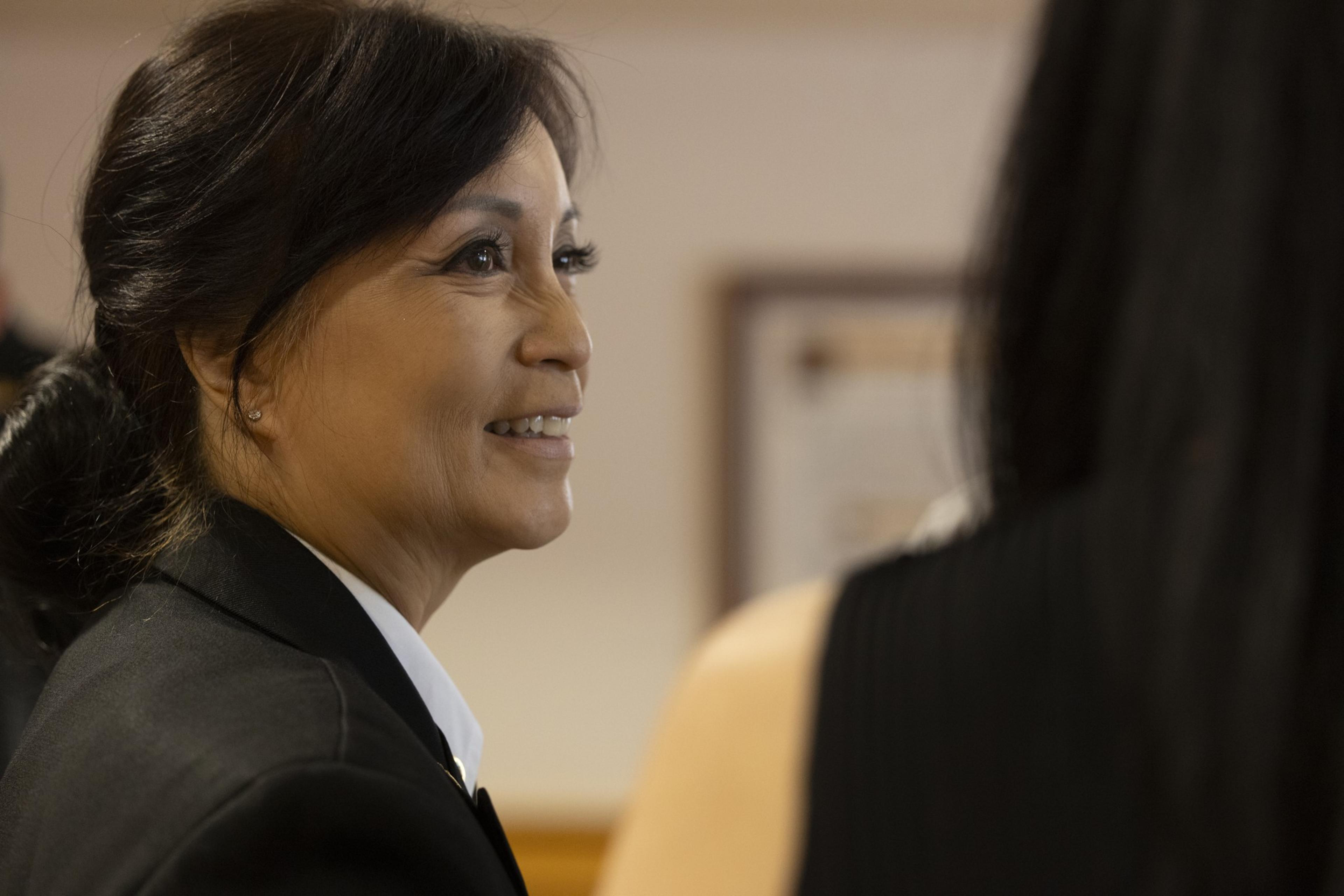 A woman in profile with dark hair tied back, wearing a black blazer and white shirt, smiles while conversing with someone who is out of focus in the foreground.