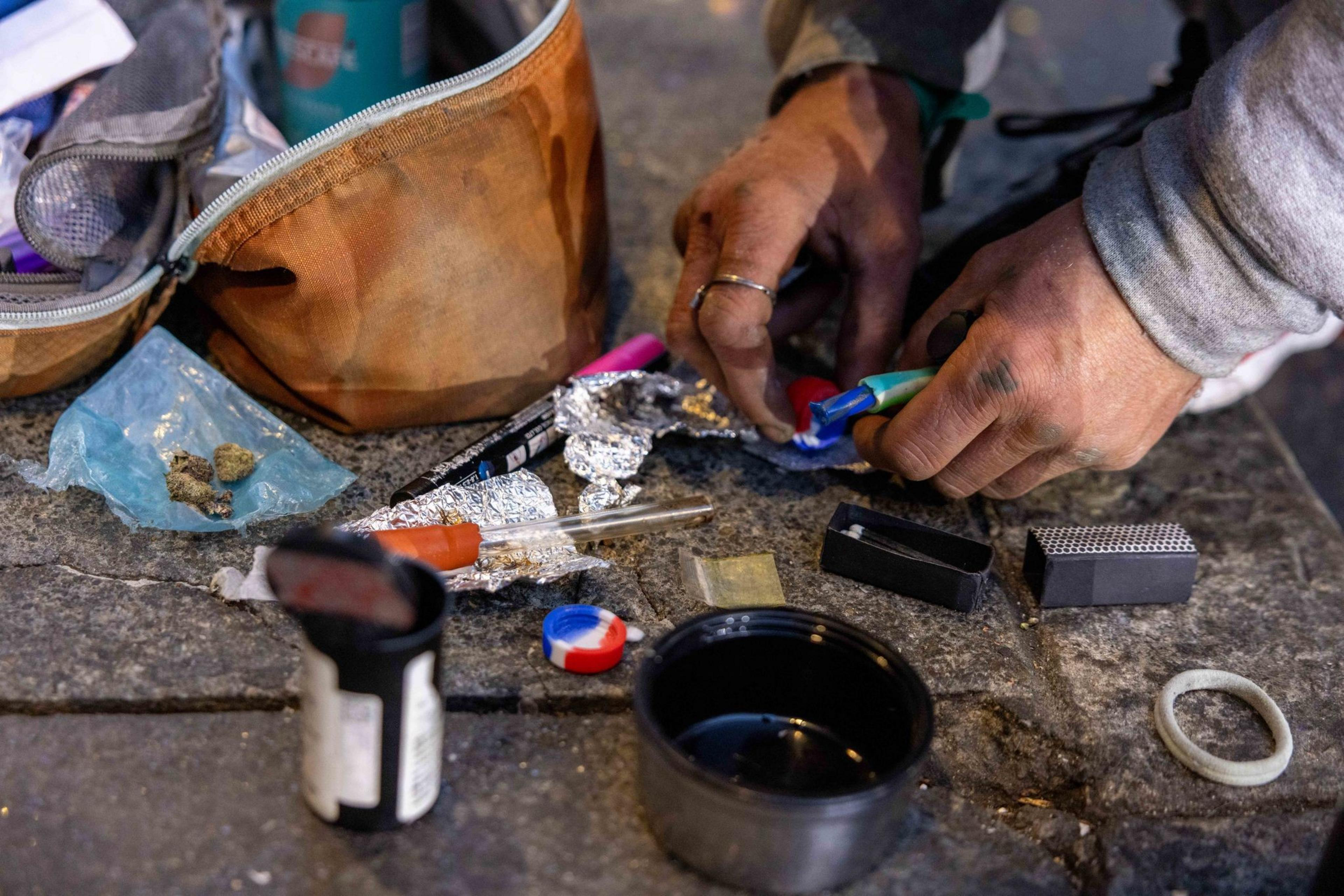 Hands are assembling drug paraphernalia on a street surface, with a pouch, foil, plastic bags, a syringe, and containers scattered around.