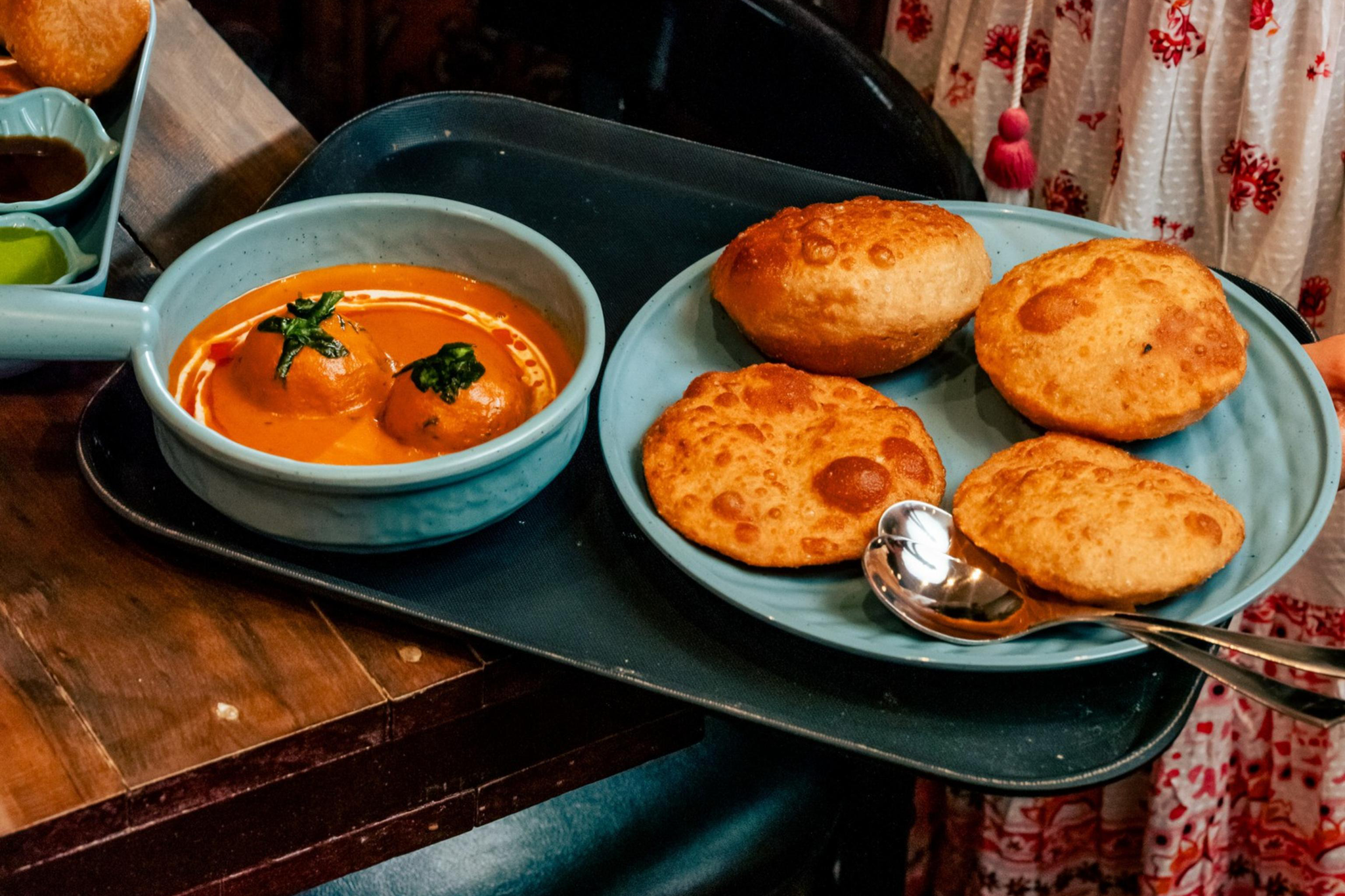 A person in a white and red floral dress holds a tray with four puffed fried bread and a bowl of curry with two round dumplings.