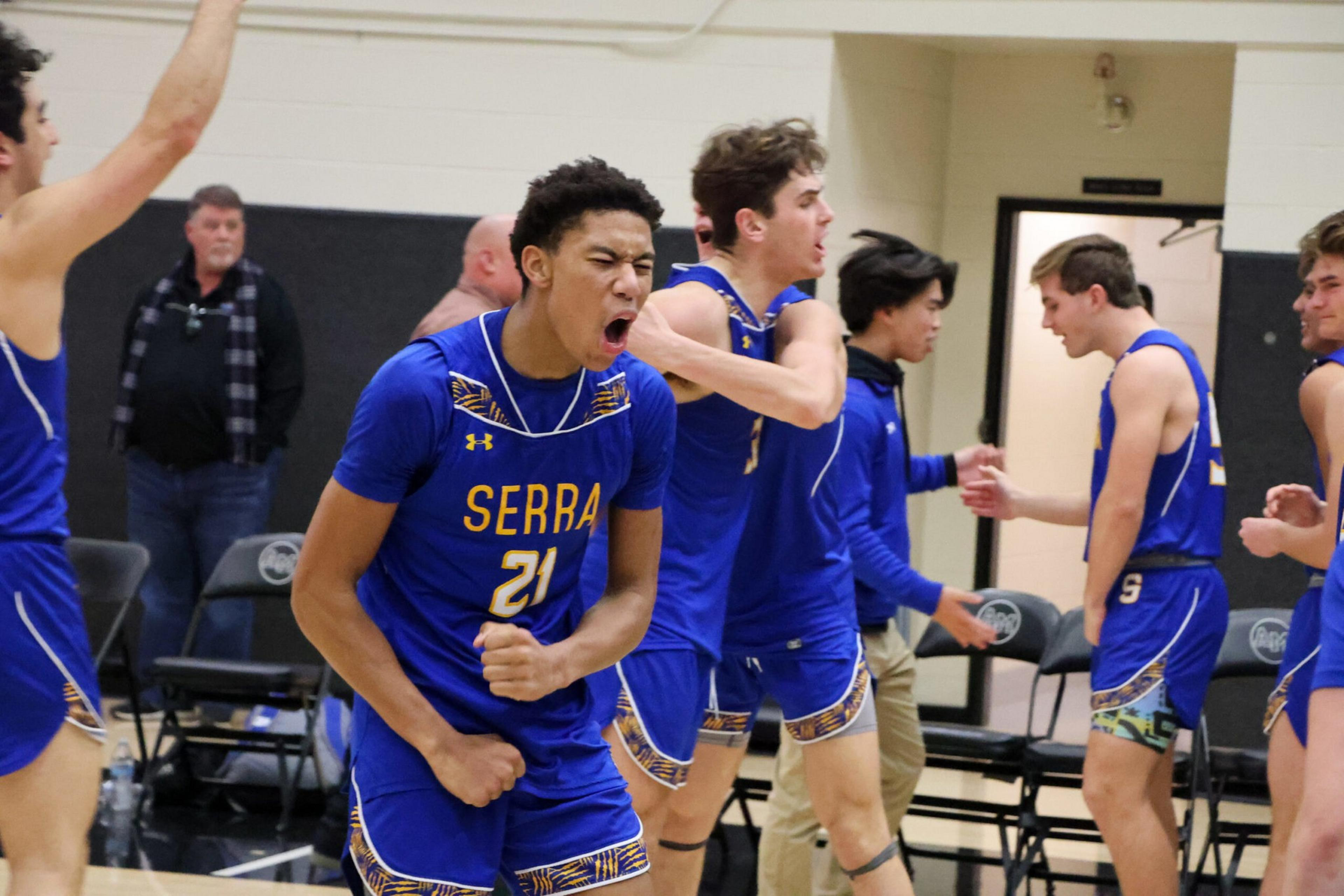 A basketball player in a blue Serra jersey yells and clenches his fists while teammates celebrate behind him.