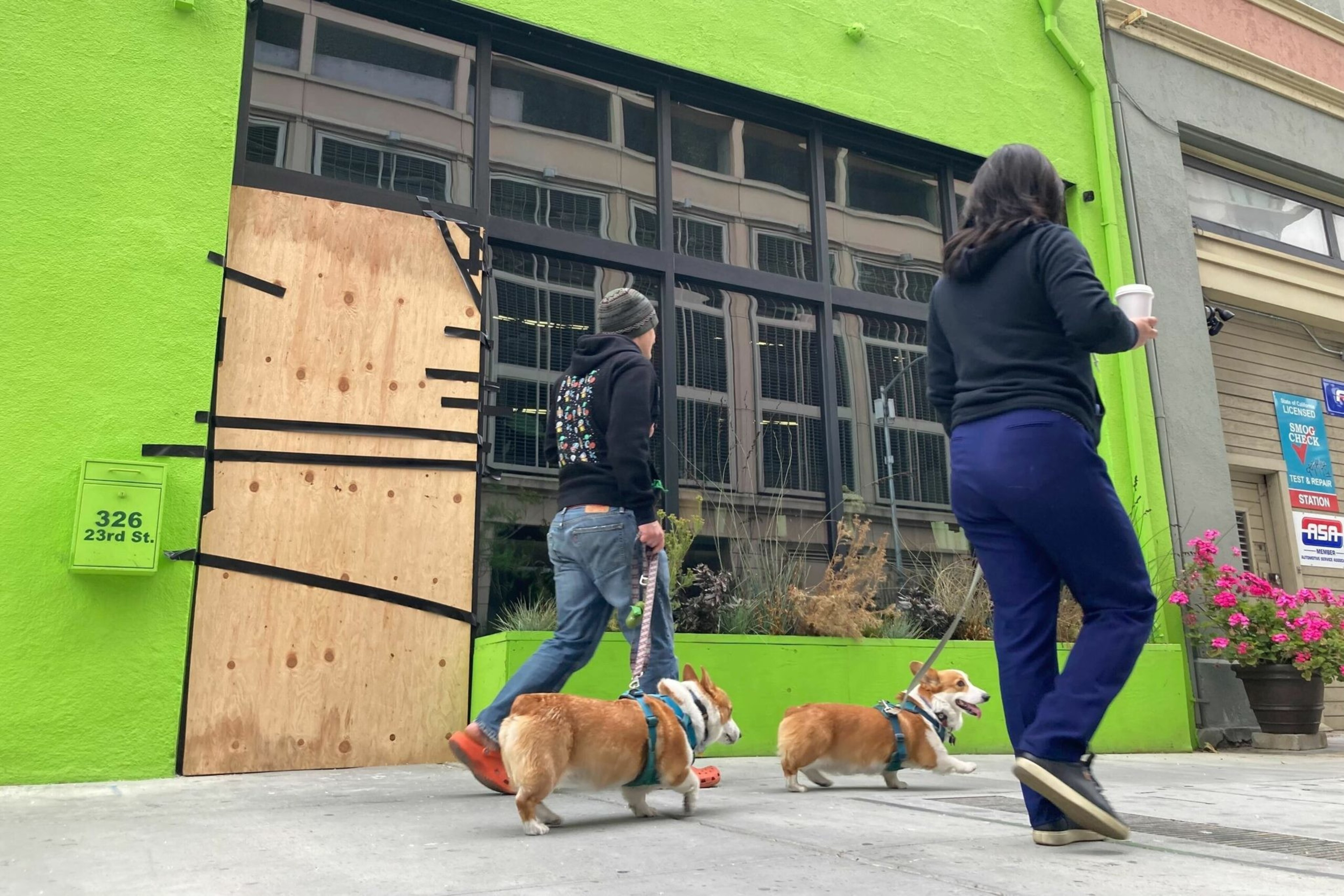 Two people walk two corgis on leashes past a bright green building with a boarded-up window and a sign that reads "326 23rd St."