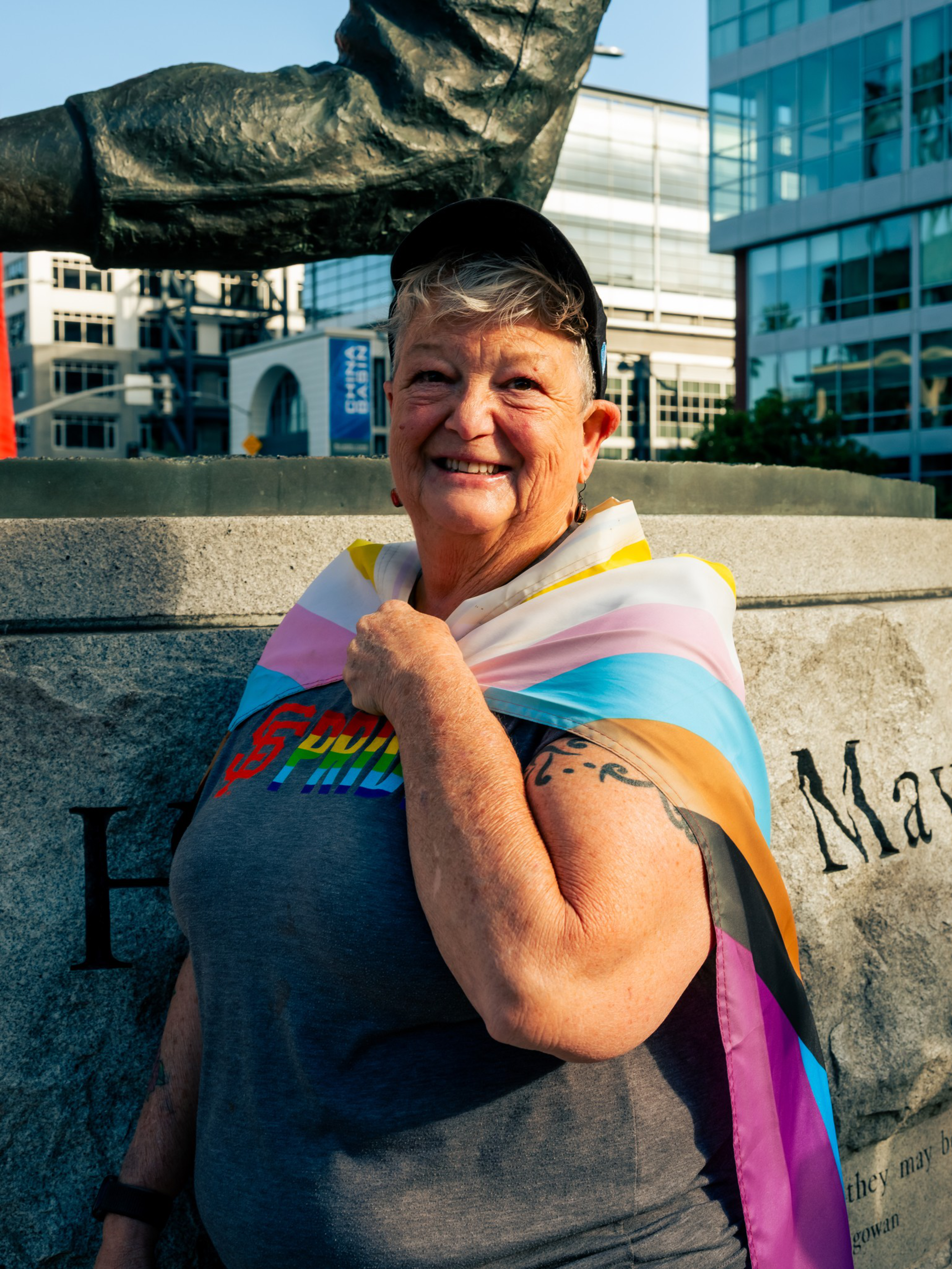 An older person with short gray hair smiles, wearing a cap, a rainbow PRIDE shirt, and a colorful pride flag draped around their shoulders.