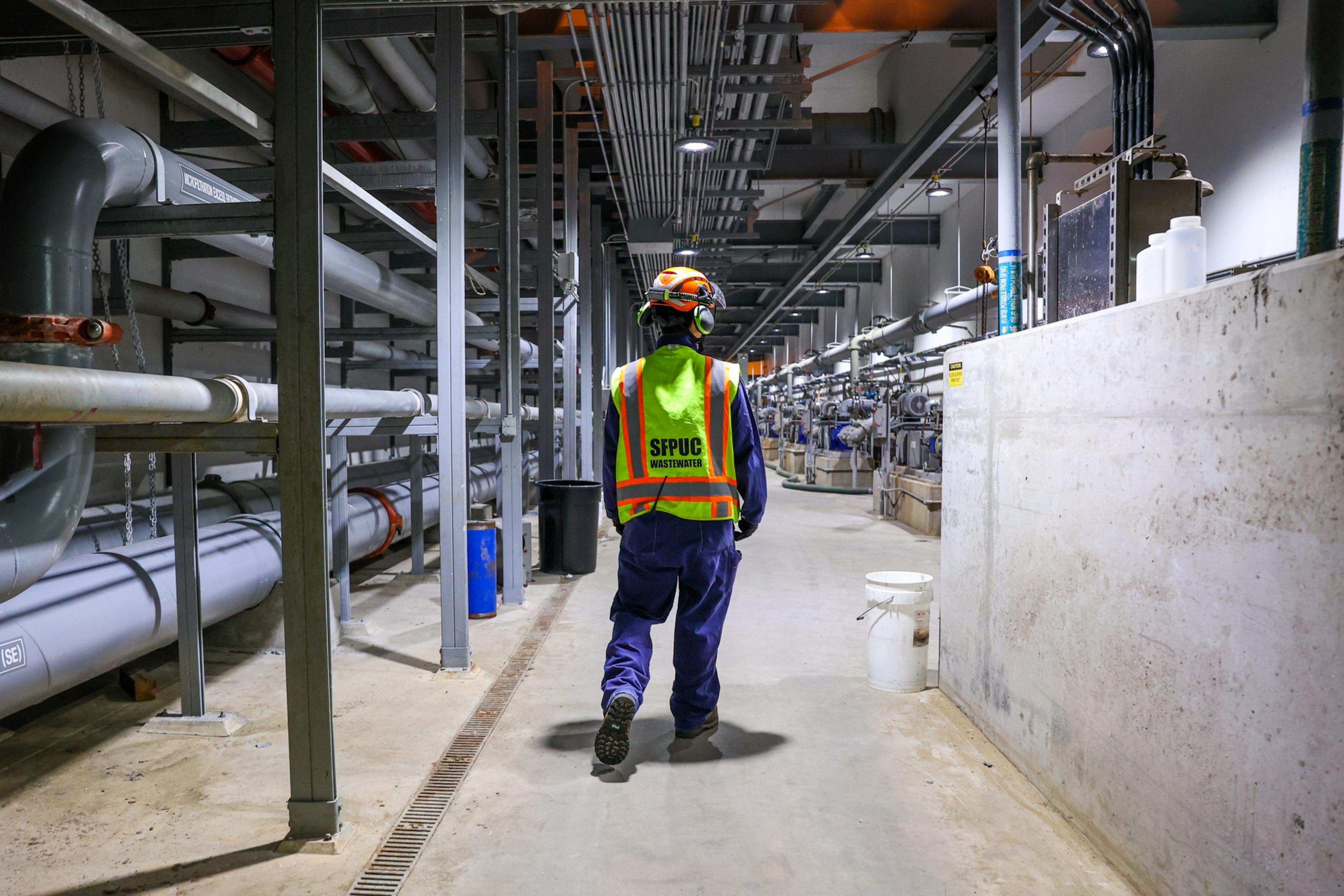 A worker in safety gear walks through a large industrial facility with pipes and machinery, wearing a vest labeled &quot;SFPUC Wastewater.&quot;