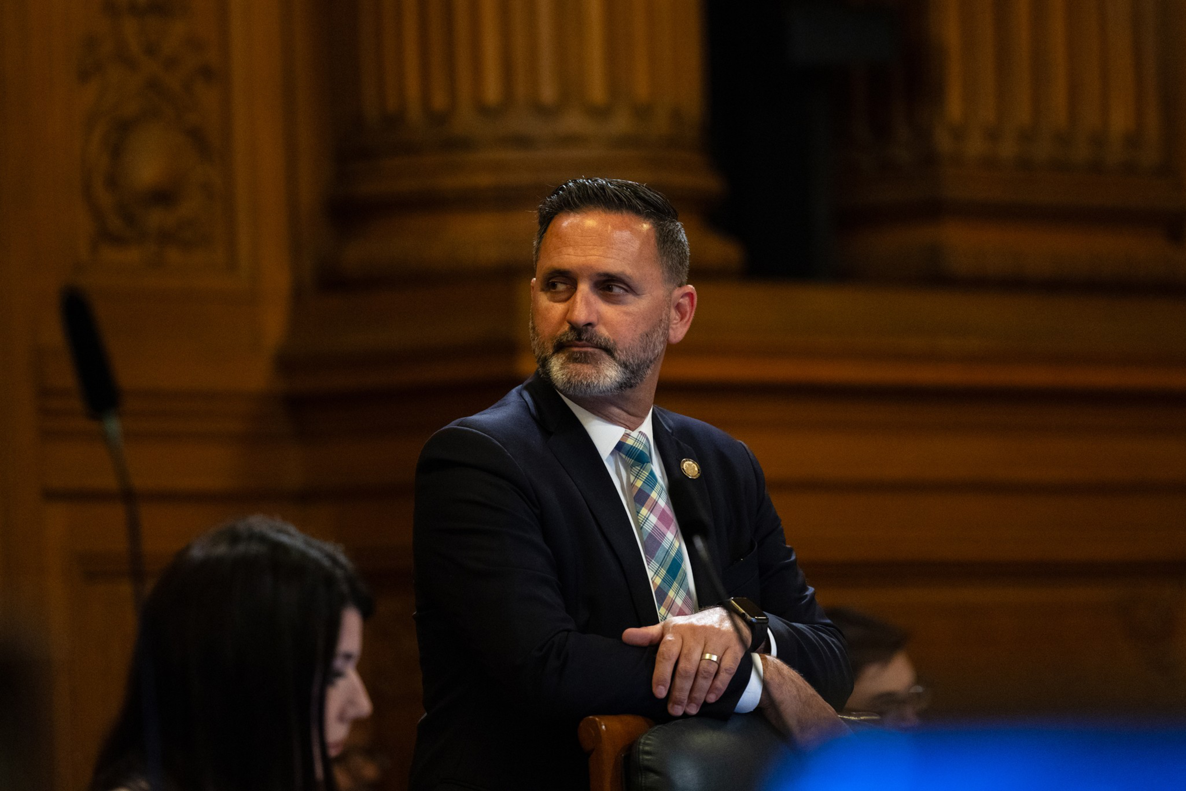 A man in a suit sits attentively, hands folded, in a chamber with ornate wood paneling.