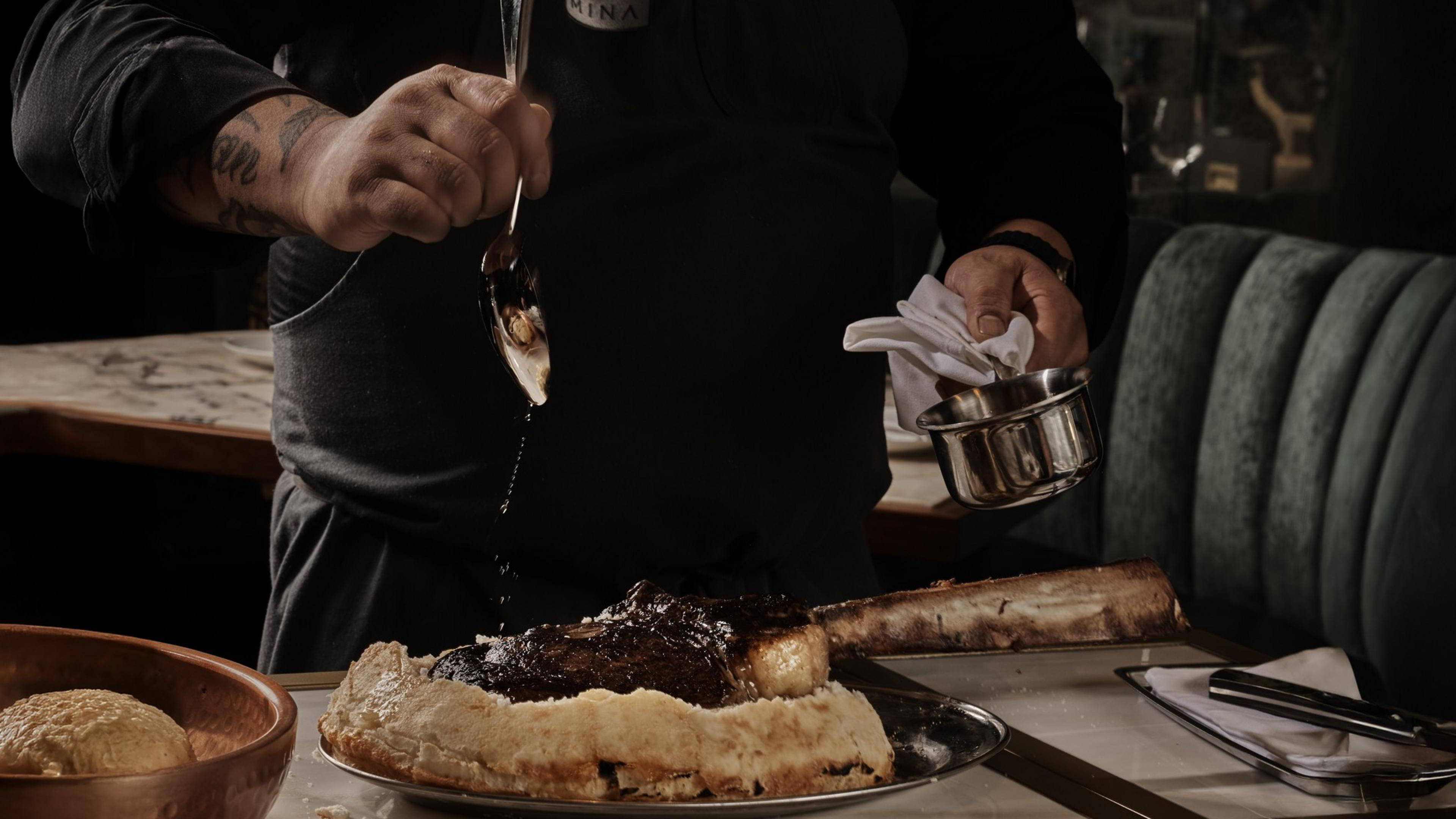 A person pours sauce over a large cooked tomahawk steak served on a fried tortilla, with a bread roll in a wooden bowl nearby.