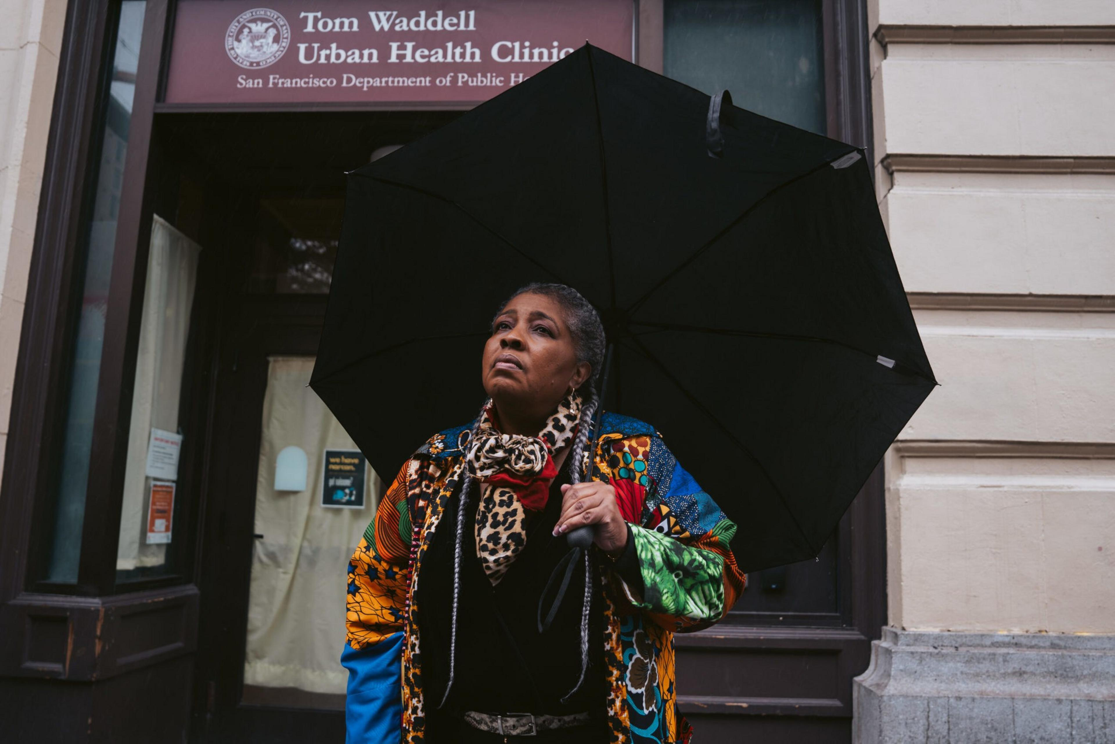 A woman with gray braided hair holds a black umbrella, wearing a colorful patterned jacket and leopard-print scarf, standing outside a health clinic entrance.