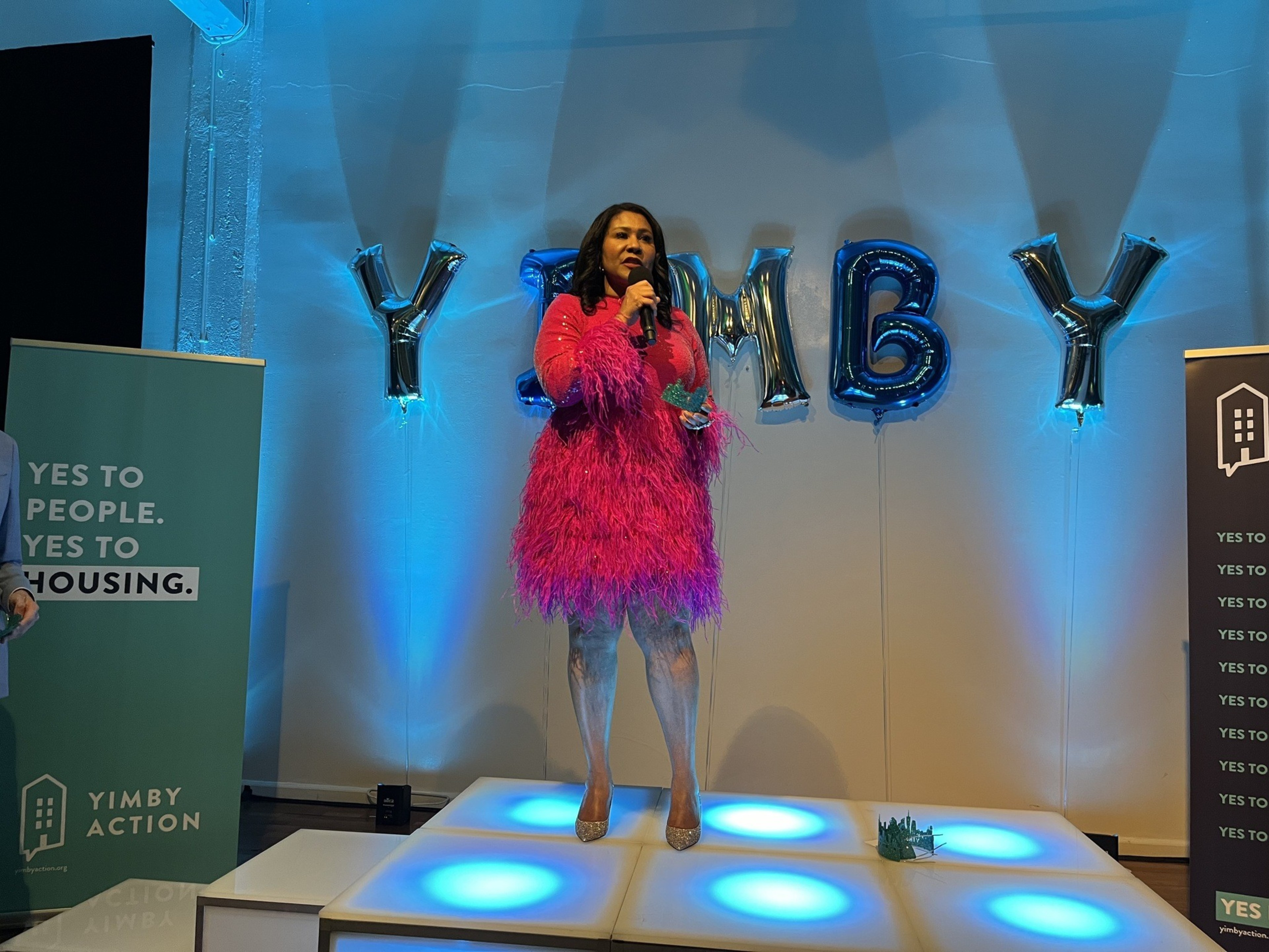 Mayor Breed in a puffy pink dress stands on a dance floor with blue lights and balloons spelling YIMBY in the background.