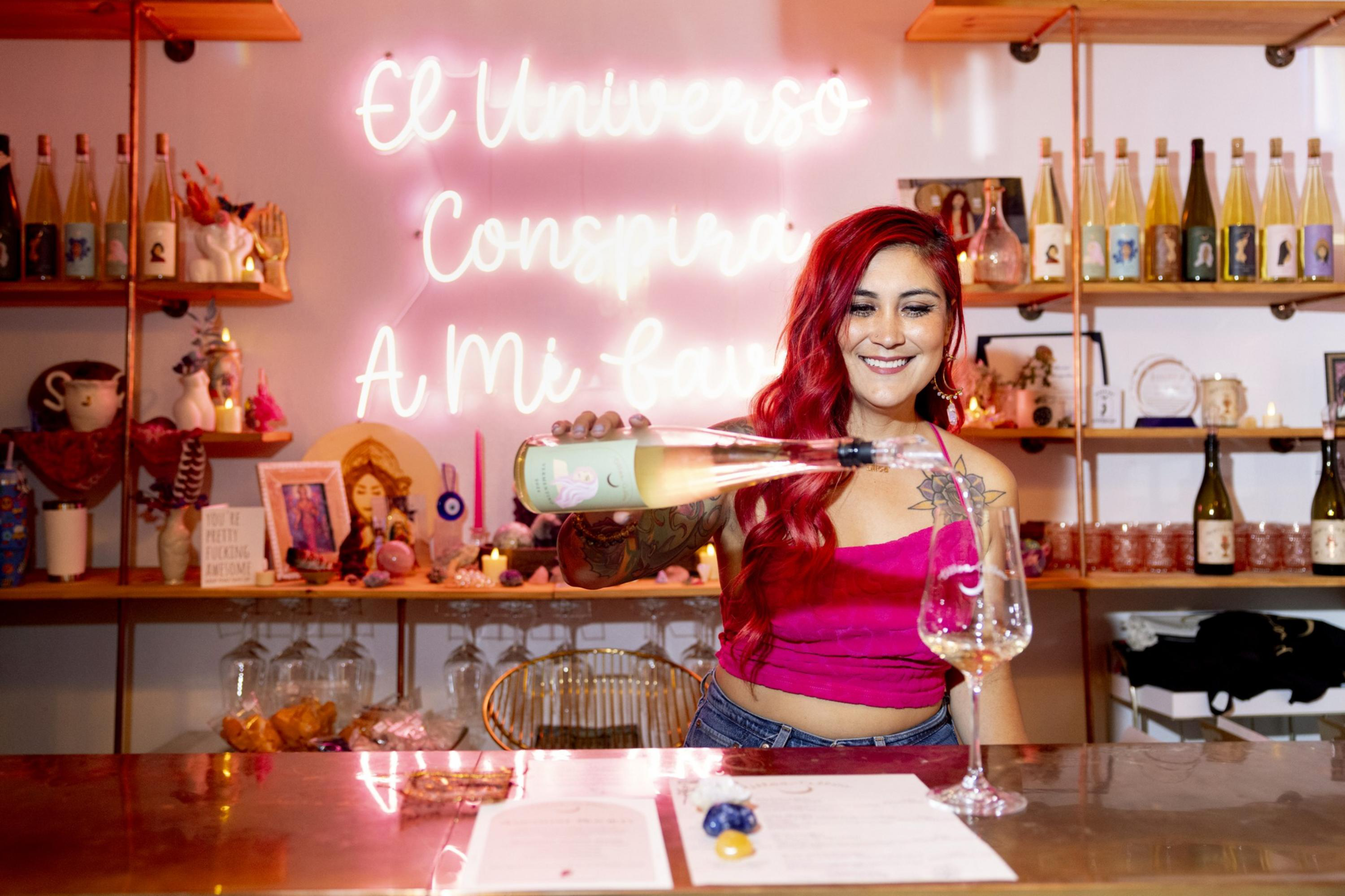 A woman with long red hair and a pink top smiles while pouring wine into a glass behind a bar with bottles and neon-lit shelves.
