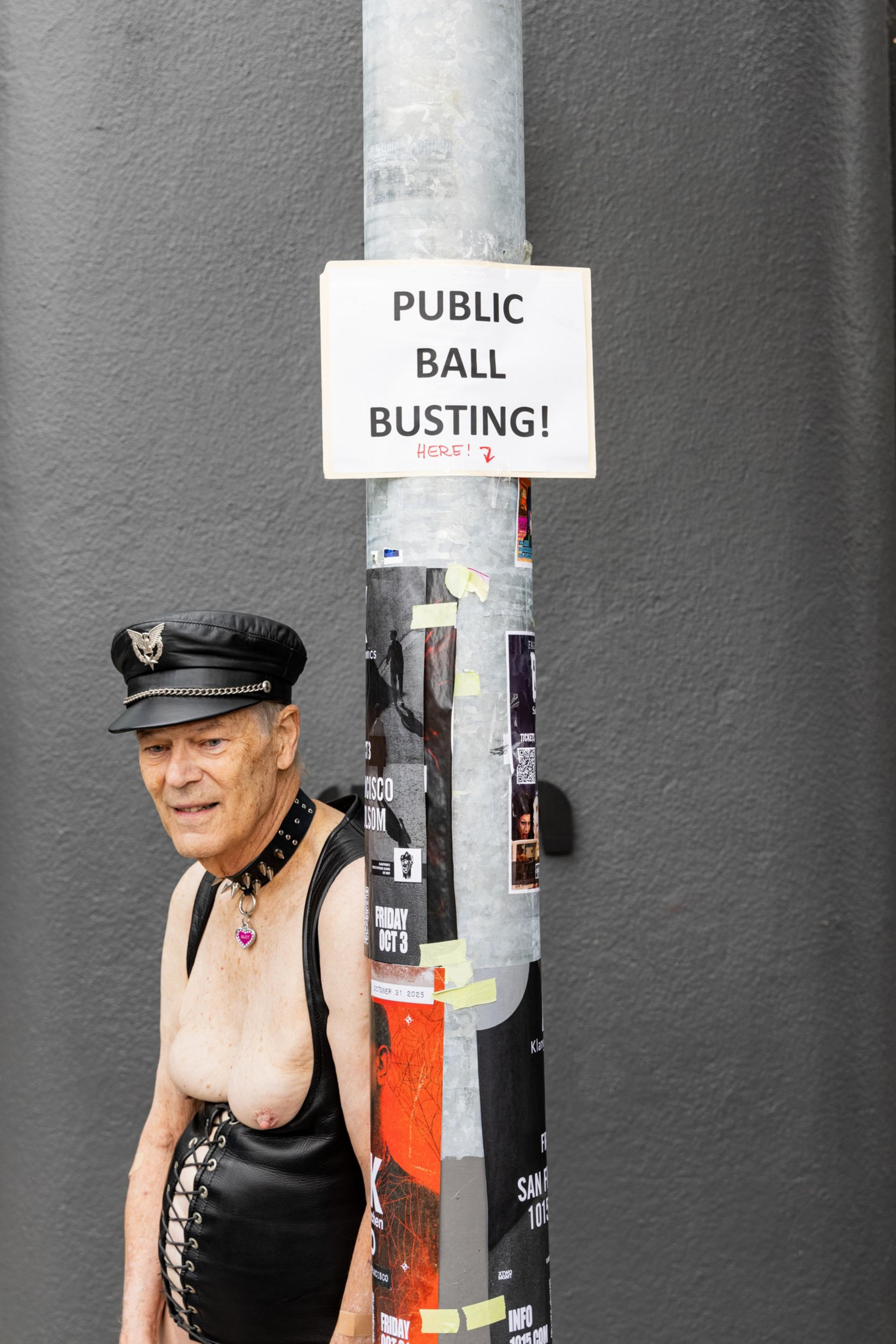 An older man wearing a leather cap, choker with a heart pendant, and a leather harness stands next to a pole with a sign that reads “Public Ball Busting! Here!”