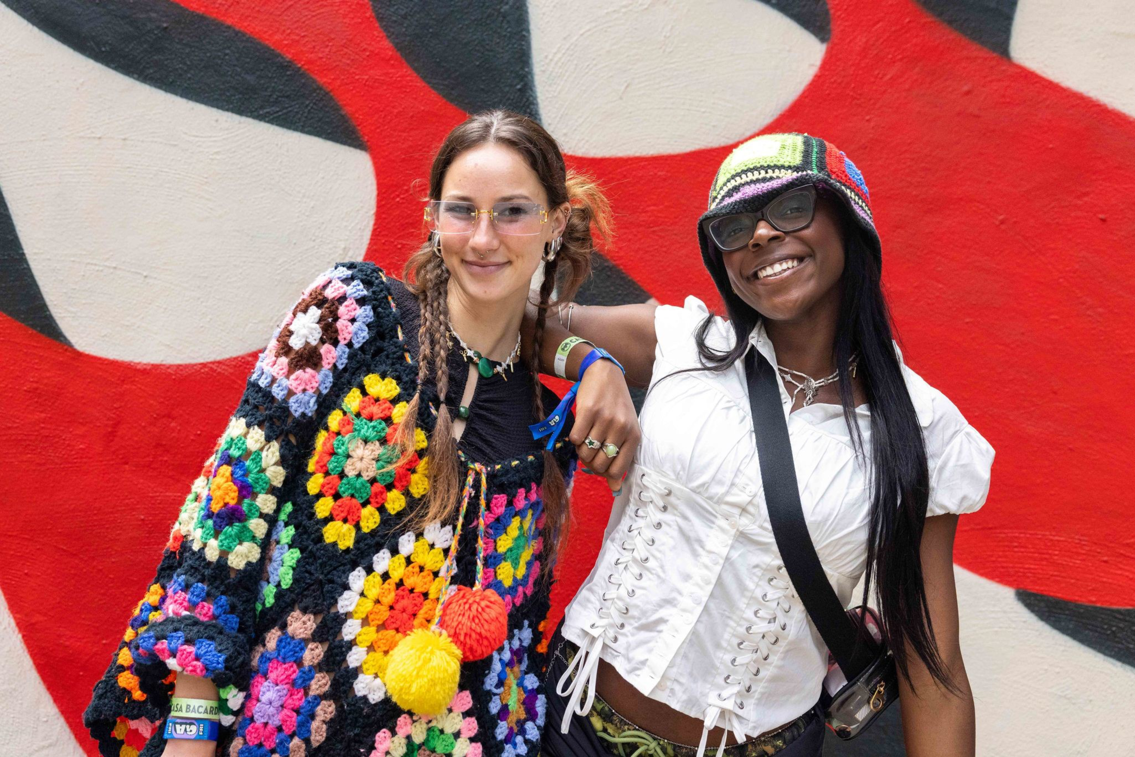 Two smiling young women stand in front of a red and white graffiti wall; one wears a colorful crochet cardigan, the other a white corset top and multicolor hat.
