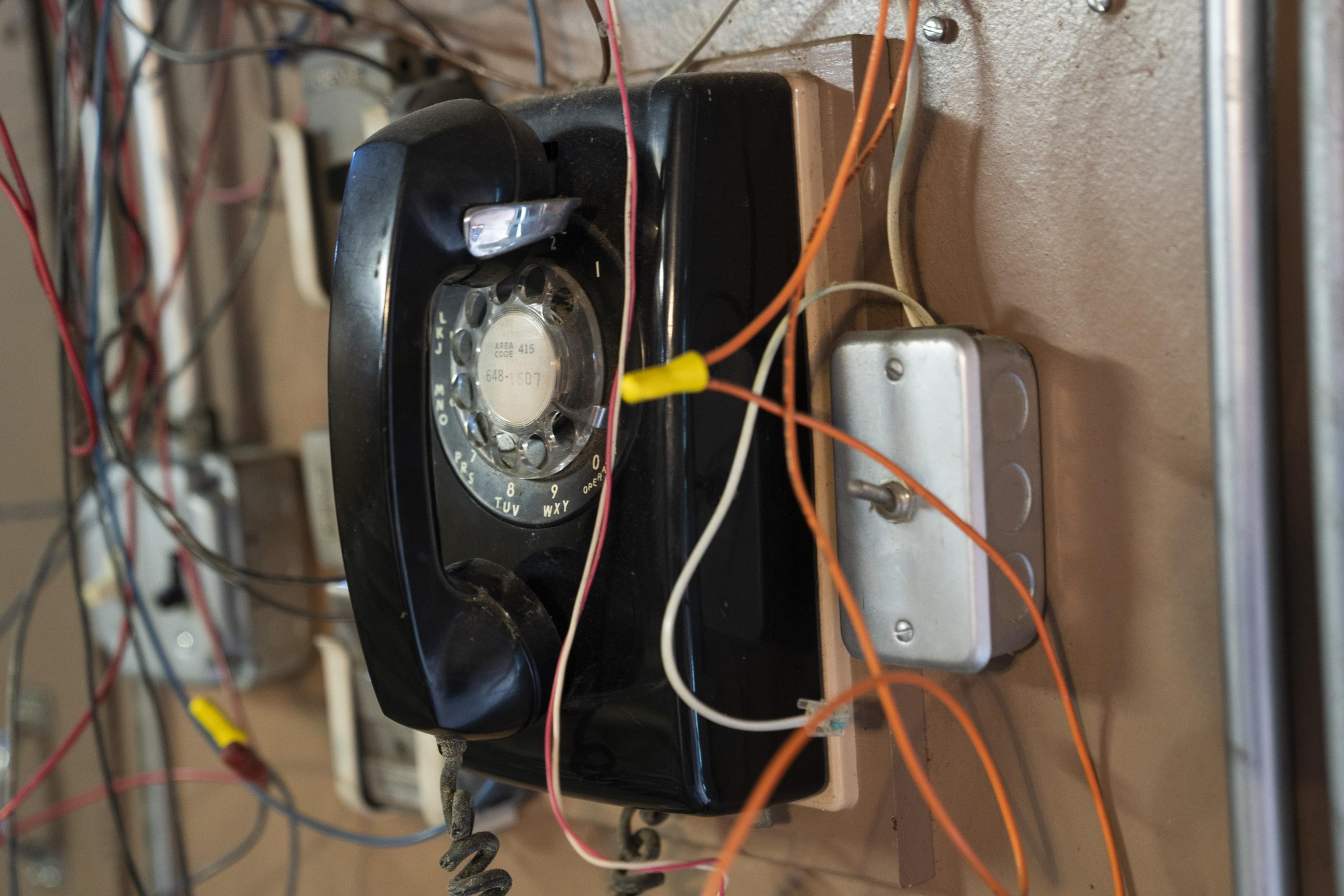 A black rotary dial telephone is mounted on a wall, surrounded by numerous tangled wires and cables.