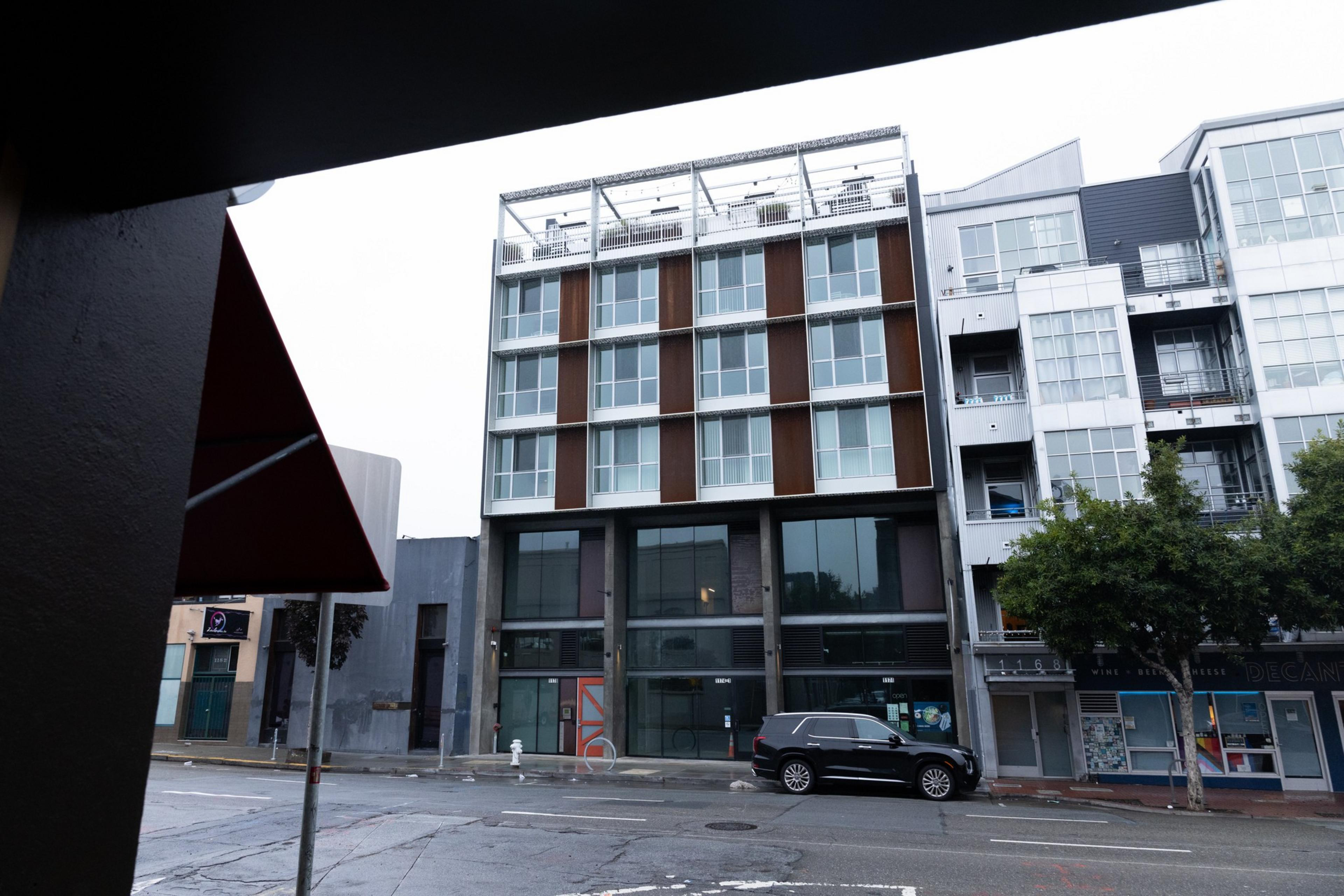 The image shows a modern, multi-story building with large windows and a mix of brown and white panels. A black SUV is parked on the street below.