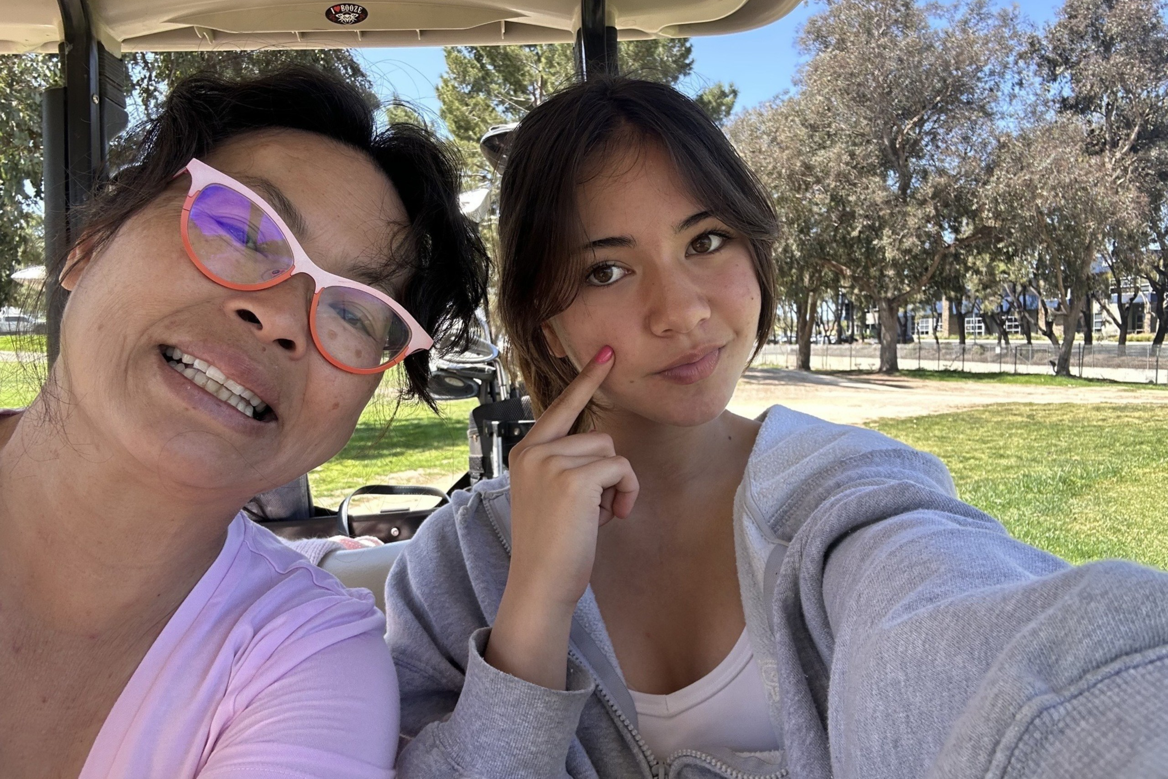Two women sit closely together in a golf cart, one smiling with pink glasses, the other touching her cheek, with green grass and trees behind them.