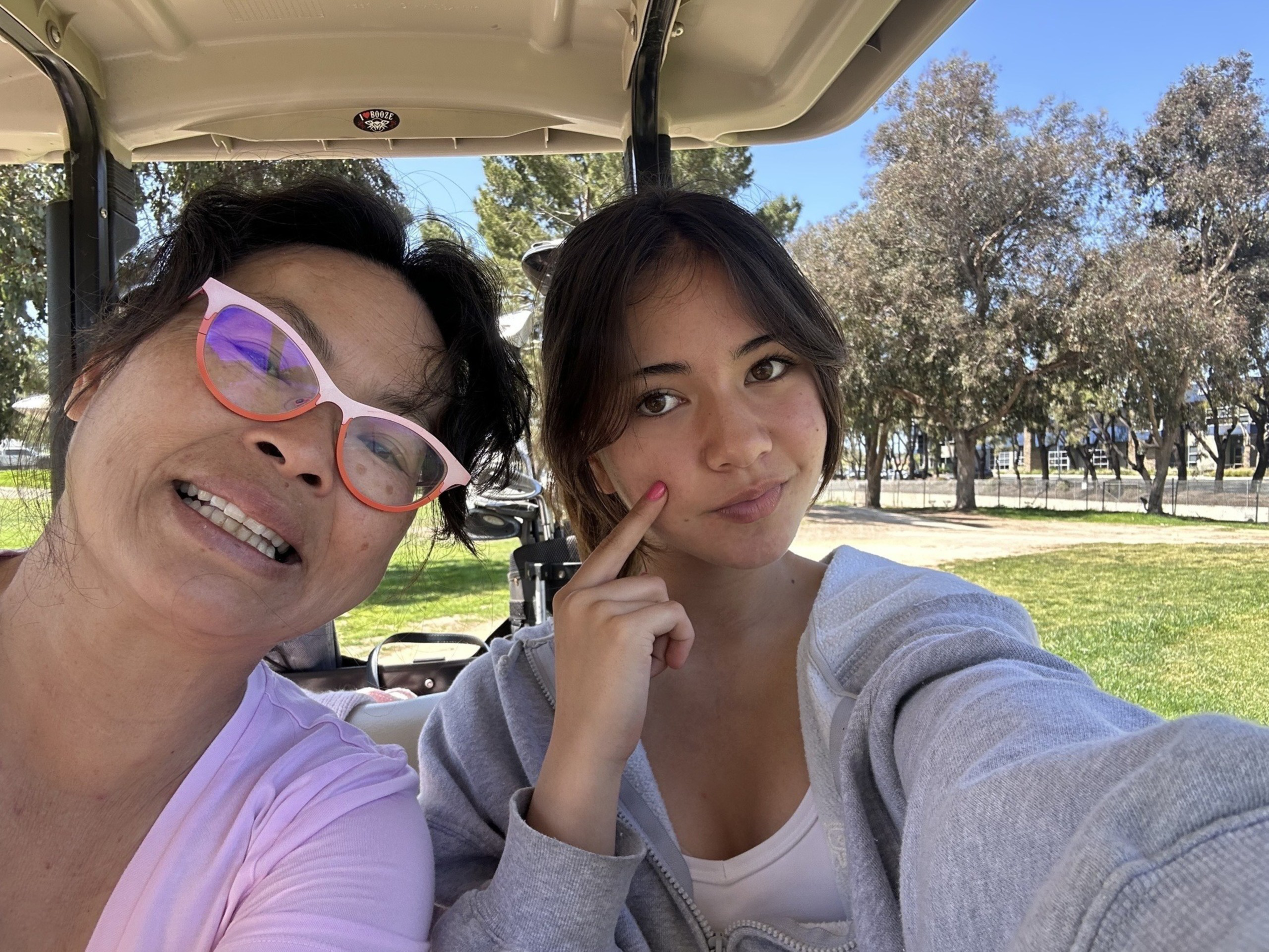 Two women sit closely together in a golf cart, one smiling with pink glasses, the other touching her cheek, with green grass and trees behind them.